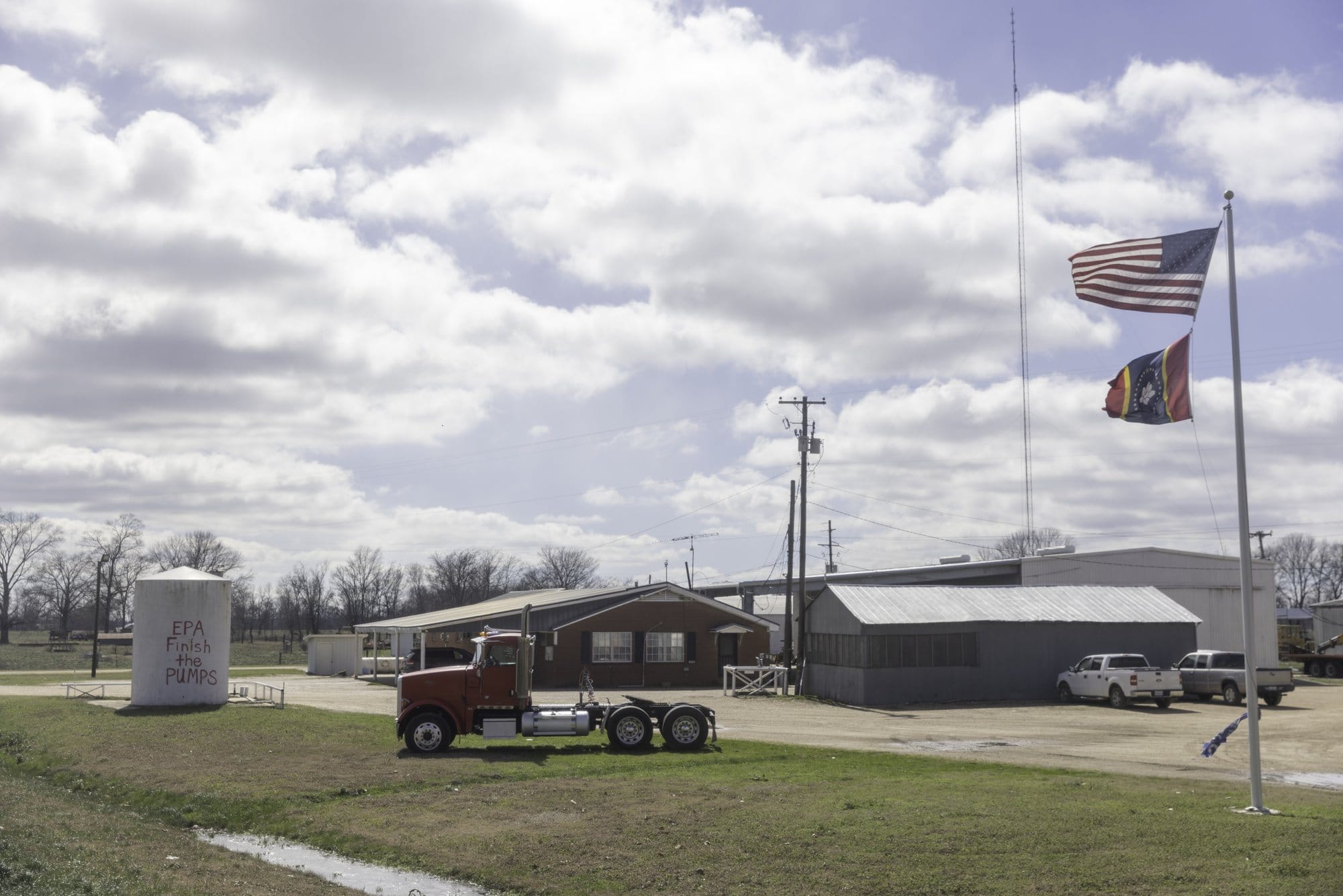 Scene of rural buildings, trucks, and waving flags, and a water tank that has been spray painted. It reads "EPA finish the pumps"