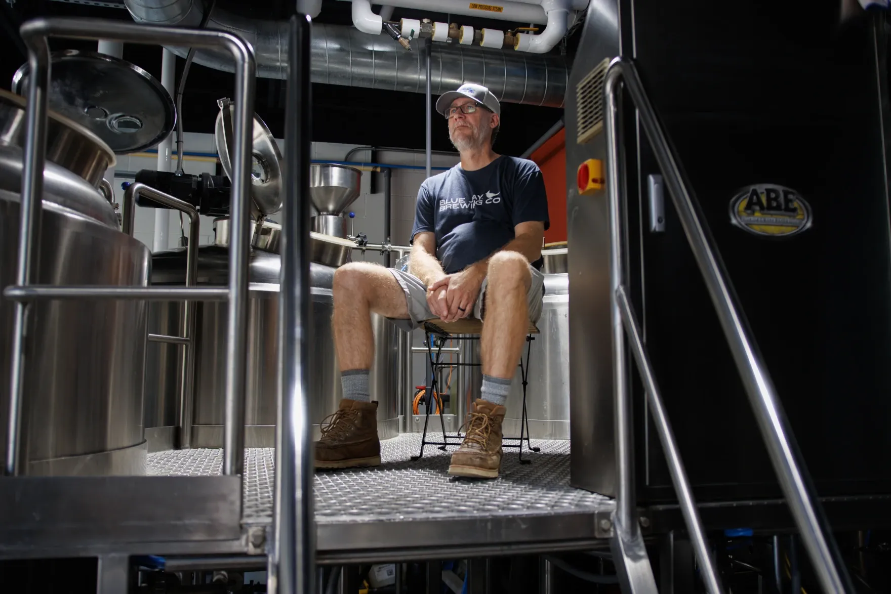 A man sits among stainless steel brewing equipment.