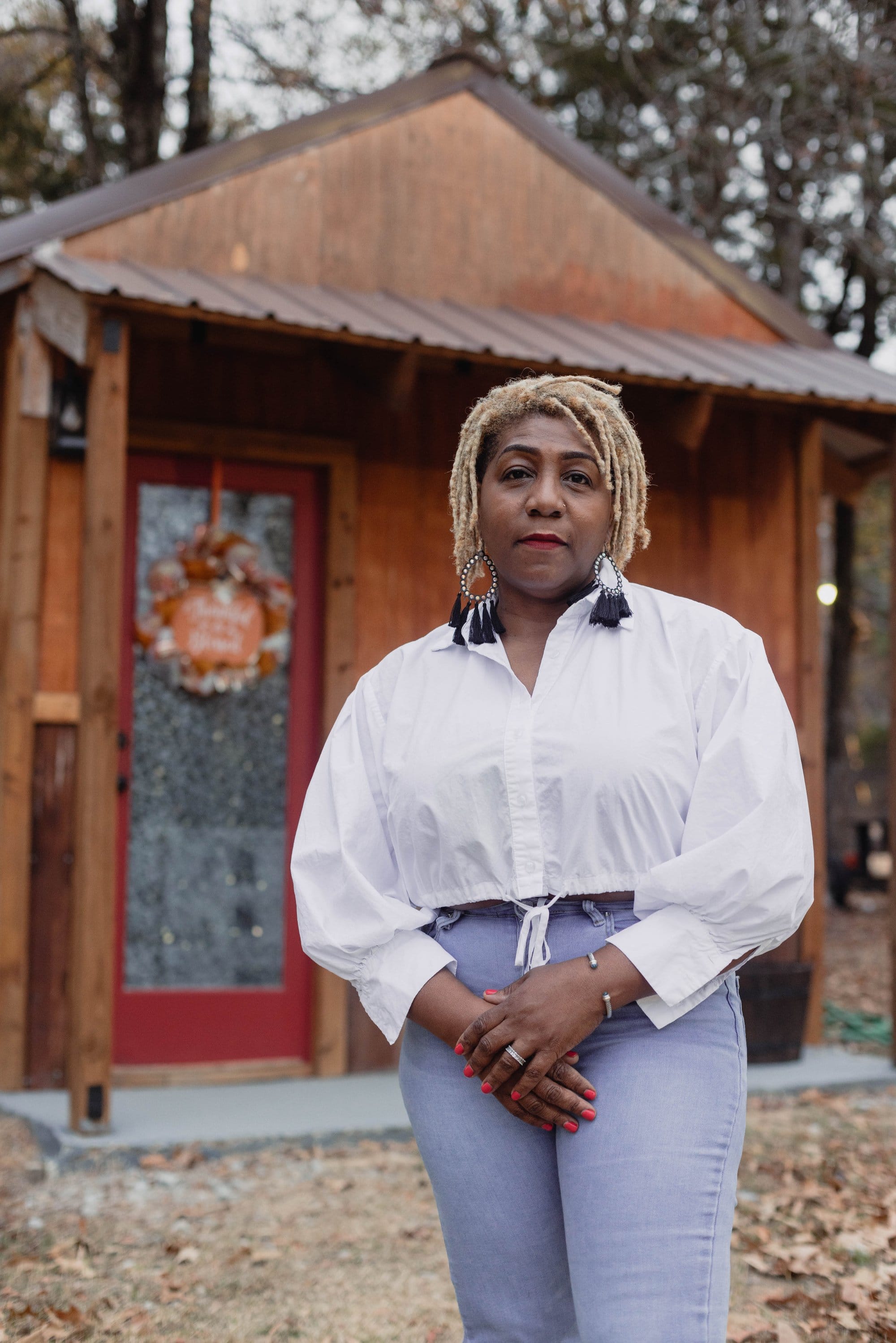A woman in a white button up shirt, blue jeans, and fringed earrings poses in front of a red-doored cabin