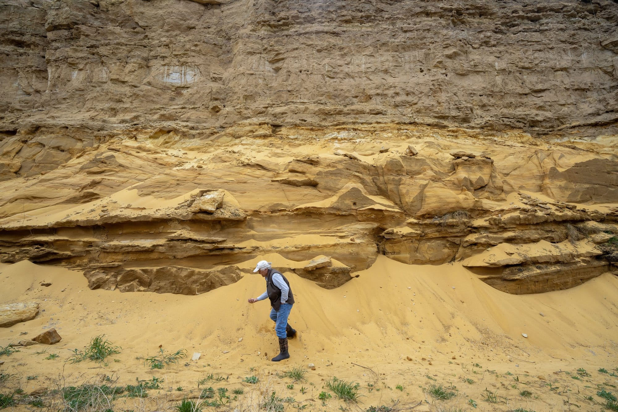 A man examines sand at the base of a layered exposed, rock face.