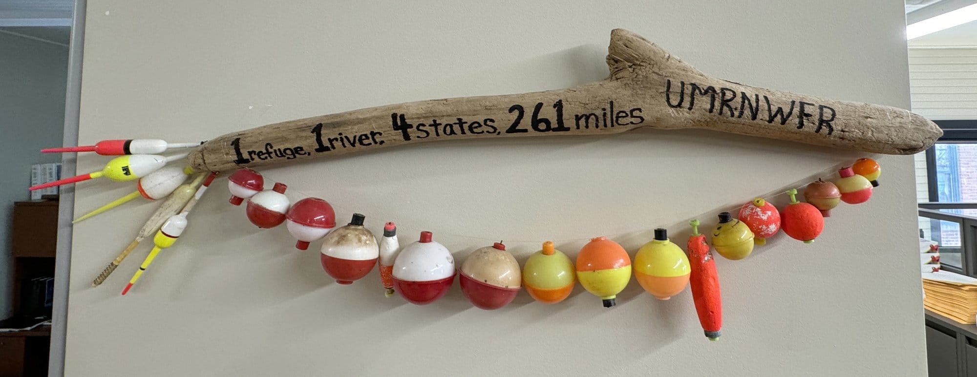 A fish-shaped installation hangs on a wall: a piece of driftwood with old round bobbers hanging down and long bobbers that make a tail. it read "1 refuge, 4 states, 261 miles, UMRNWFR