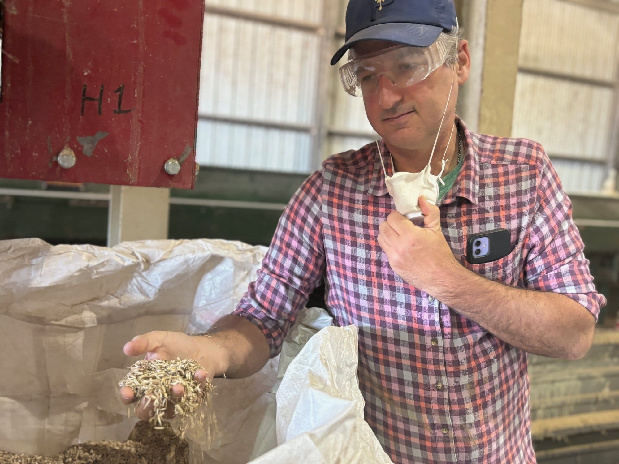 A man in safety glasses and a ballcap pulls an N95 mask down as he shows a handful of seeds
