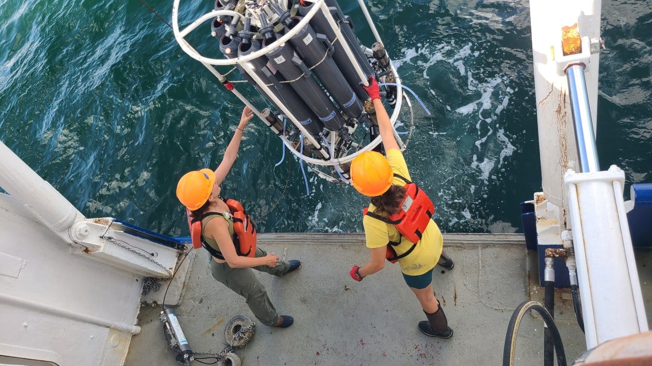 Two scientists in hard hats and life vests grab for a piece of equipment that is parially submerged in water