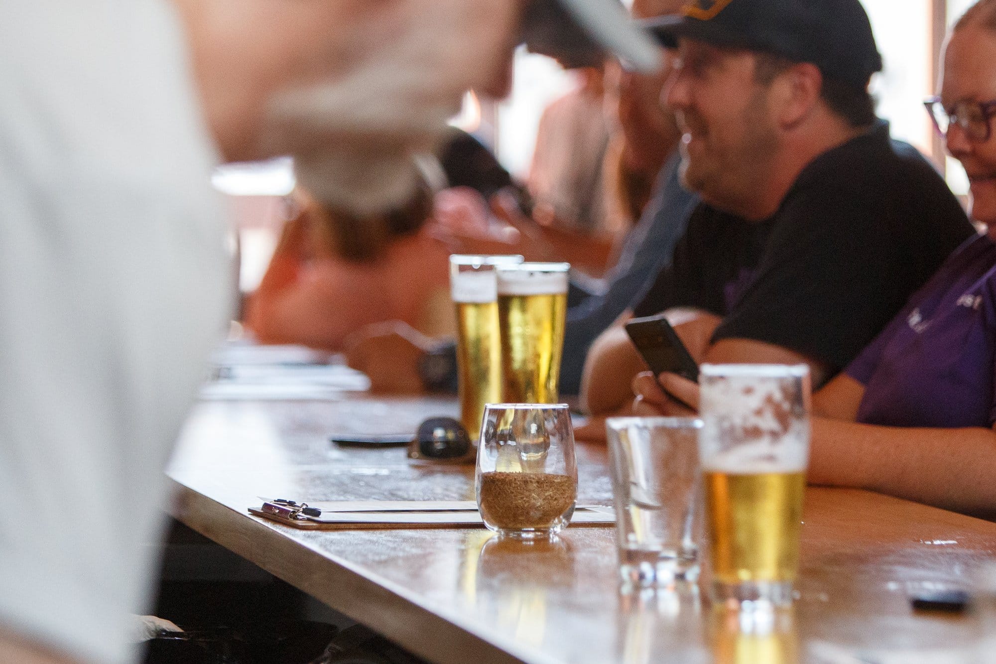 Several glasses of Kerns of beer sit on a bar as patrons gather around.