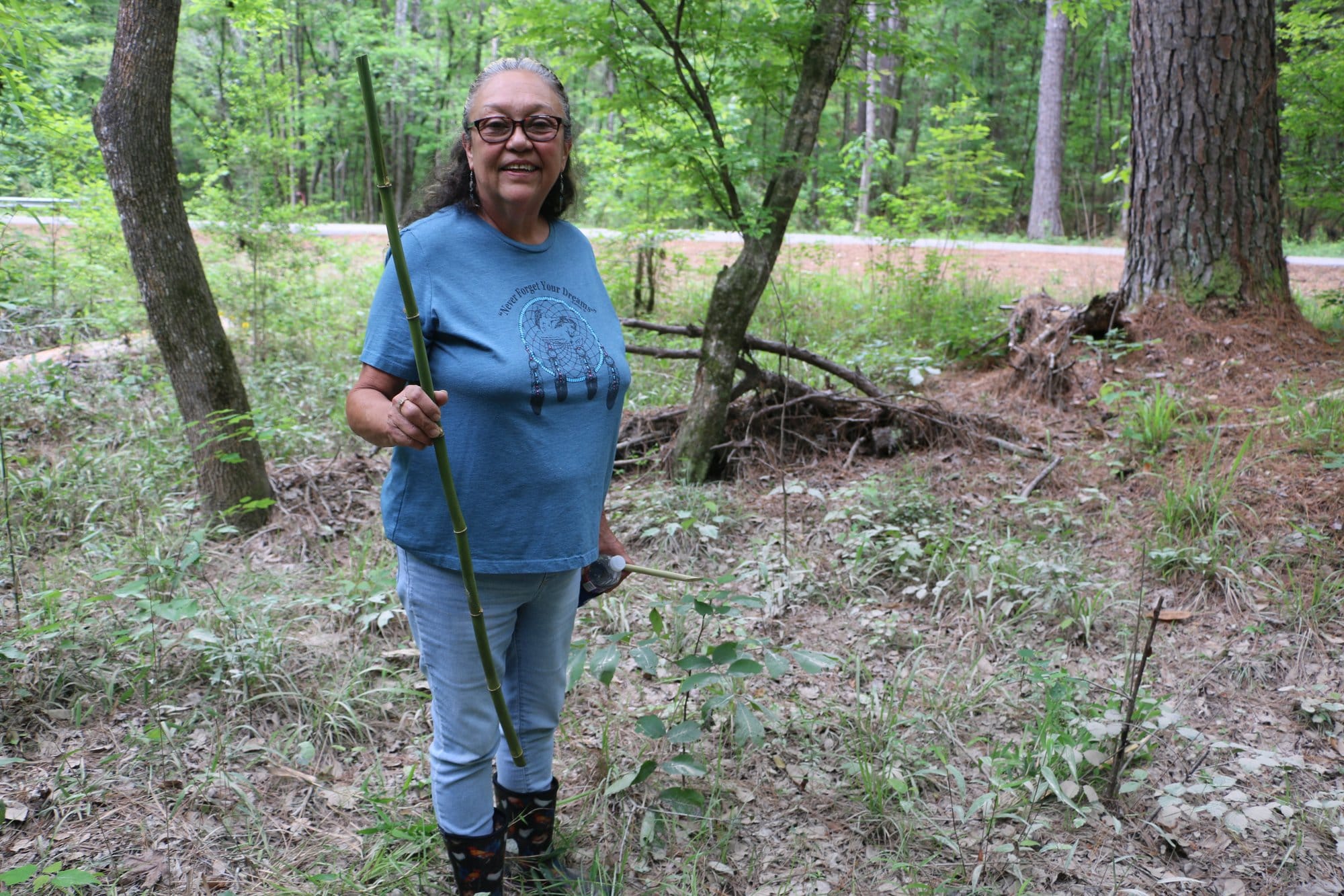 Rose Fisher Greer stands in a wooded area holding a stalk of river cane she harvested for traditional basket making.