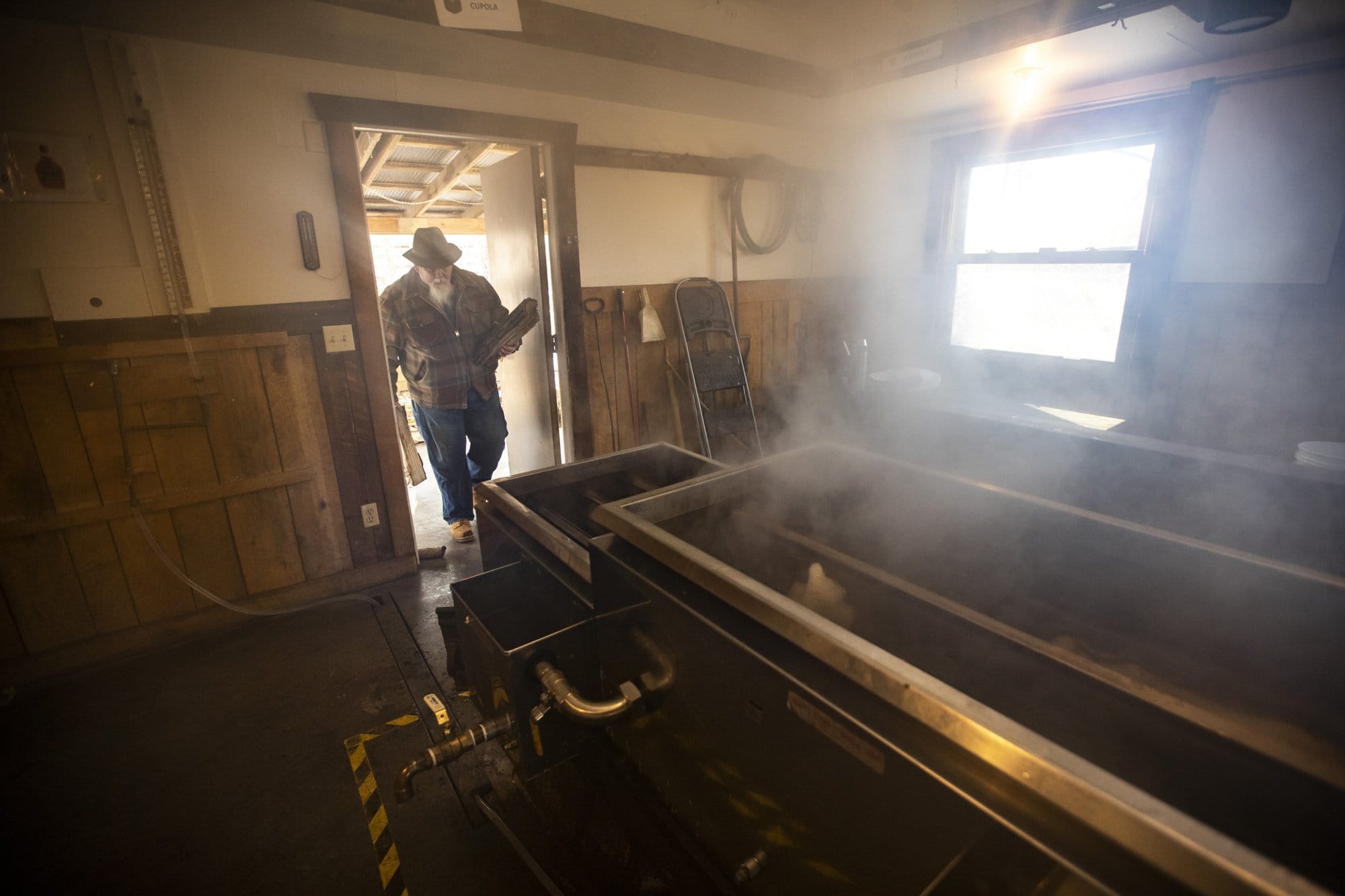 A man in a beard, flannel, and cowboy hat carries lumbeer into a small cabin room with a piece of smoking black equipment