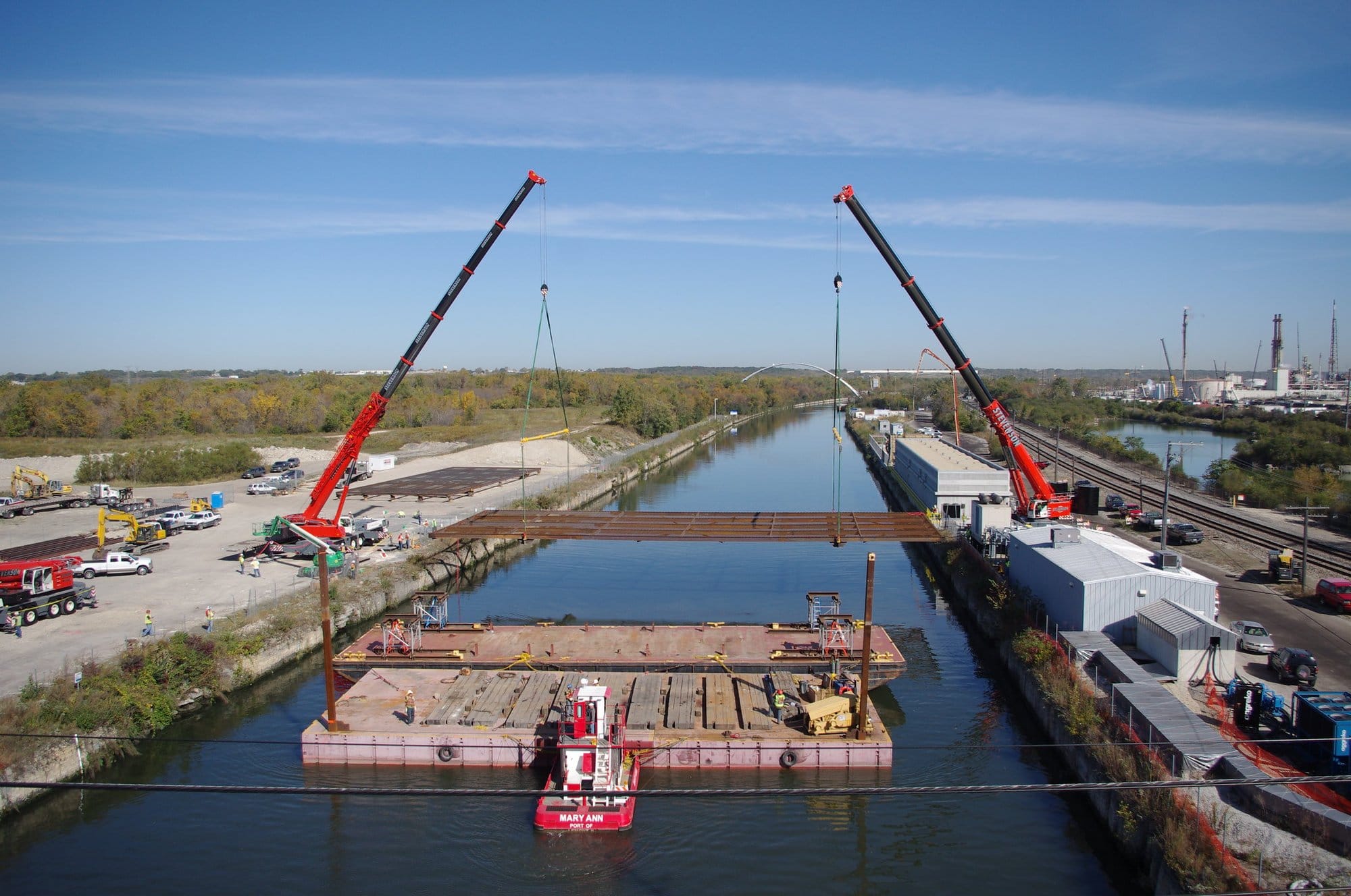 A construction crew works from either side of a river. In the water is a flat deck. Two long equipment arms lower a piece of metal onto the decks.