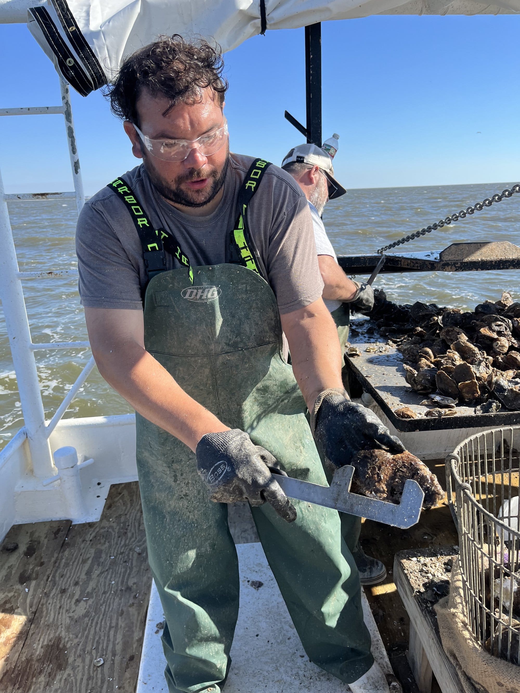 A man in a wetsuit uses a handheld tool to work with an oyster. Behind him, another man works with a table full of oysters