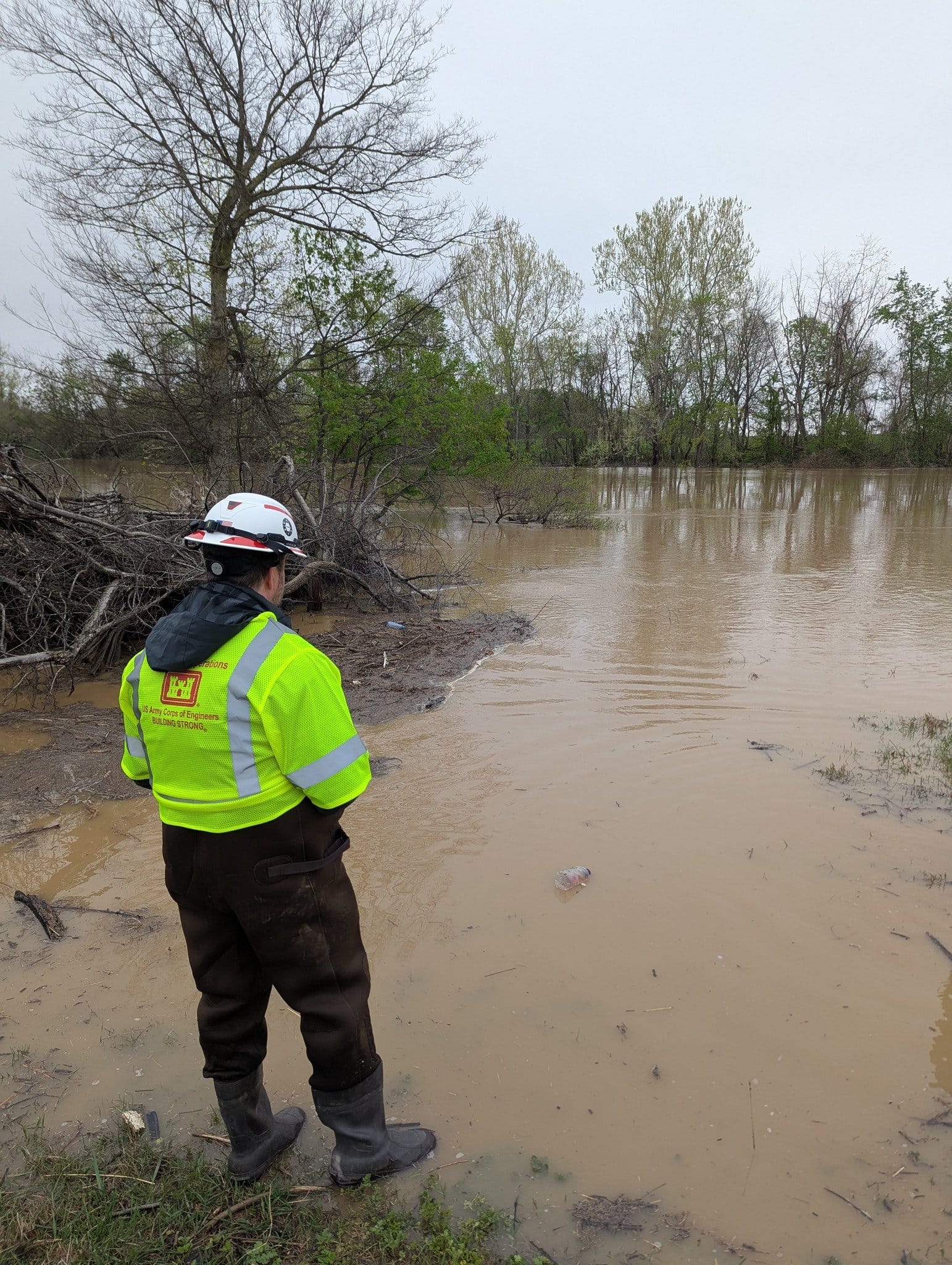 A man in a yellow safety jacket and hard hat surveys a flooded, brown body of water