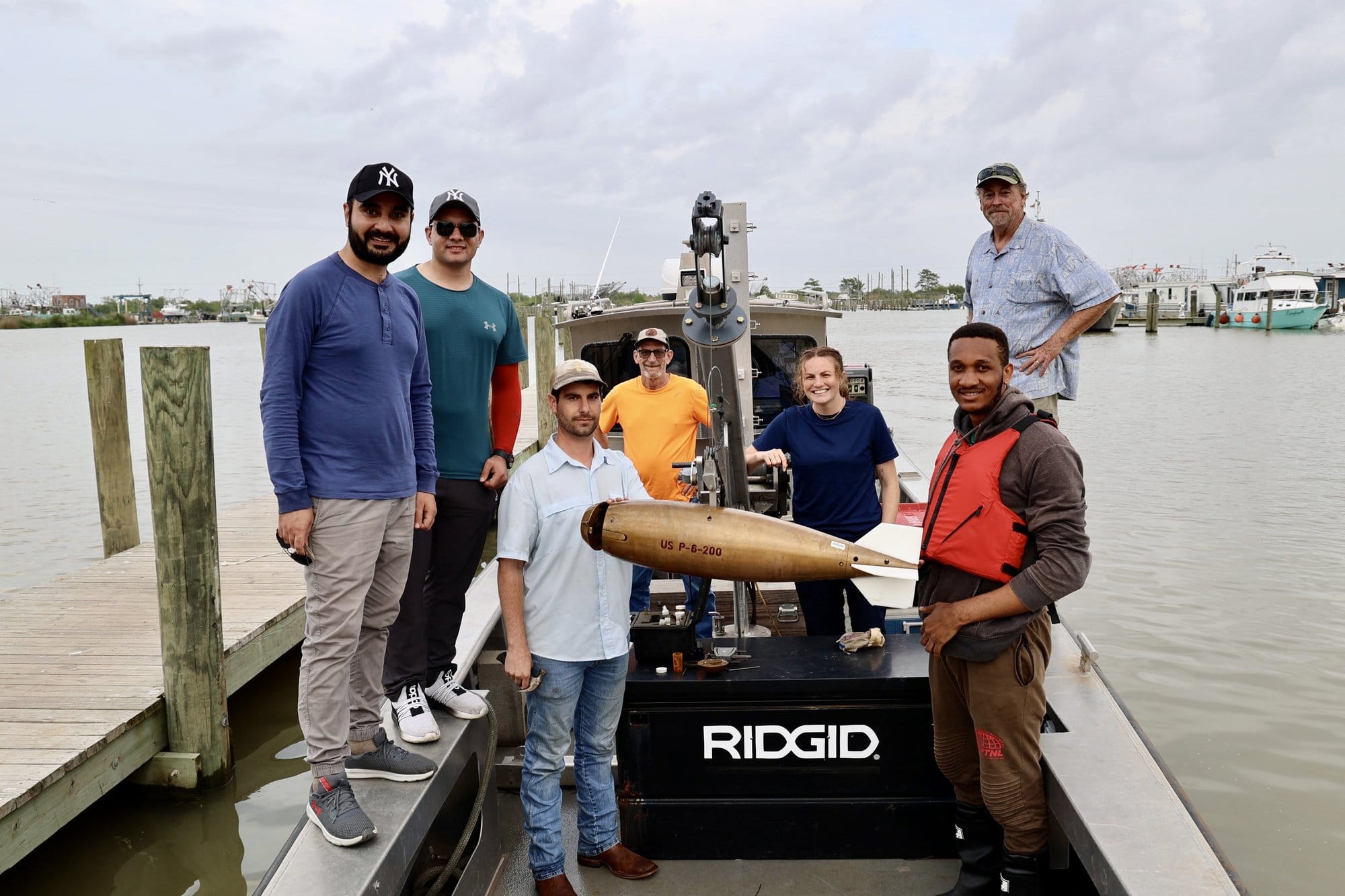 A group of people pose on a docked boat with a gold instrument that is held on heavy-duty retractable wire