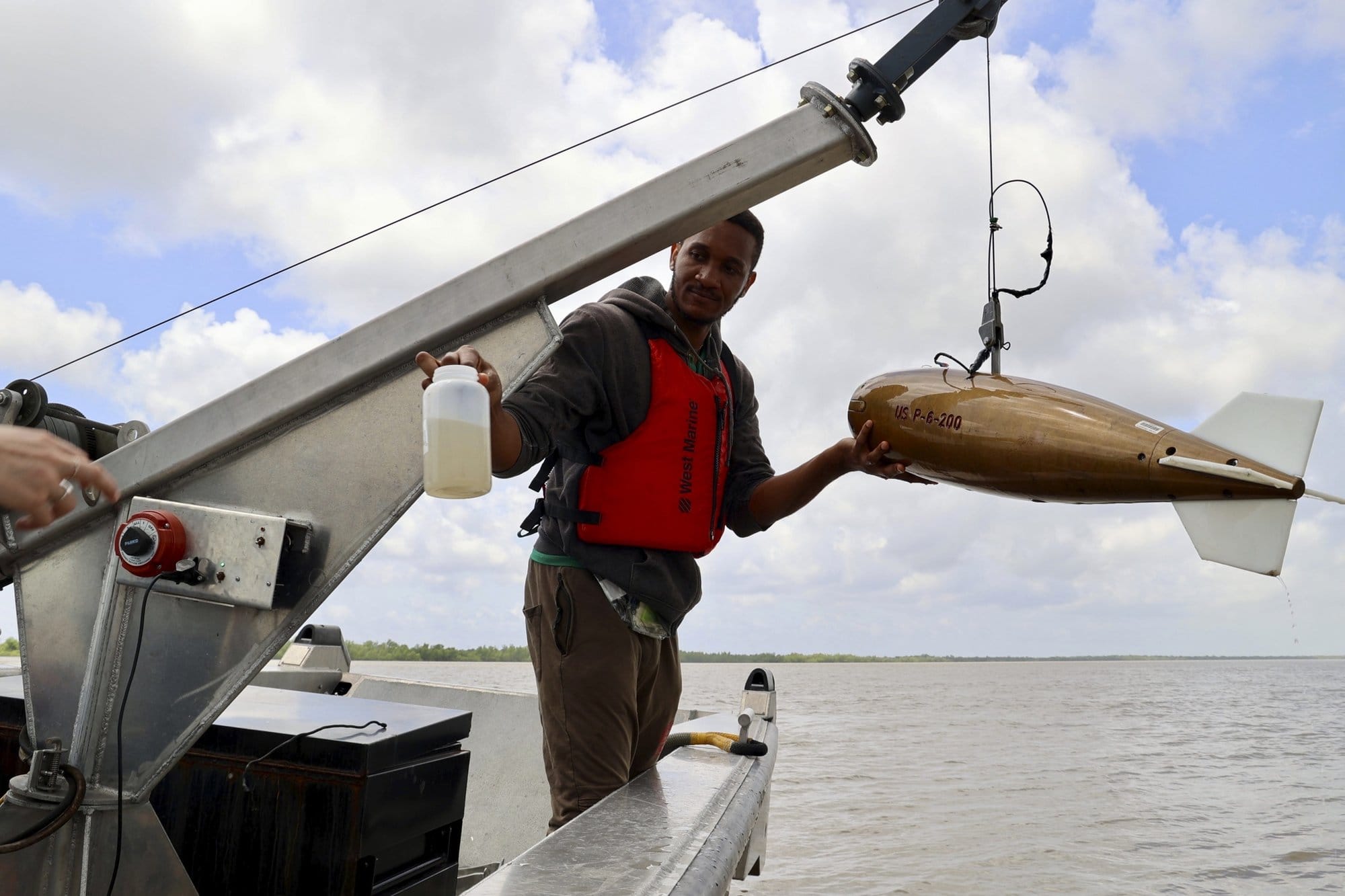 A man in an orange life stands with a large gold fish-shaped piece of equipment that hovers over the water from the edge of a boat. He is holding a lab bottle containing murky water 