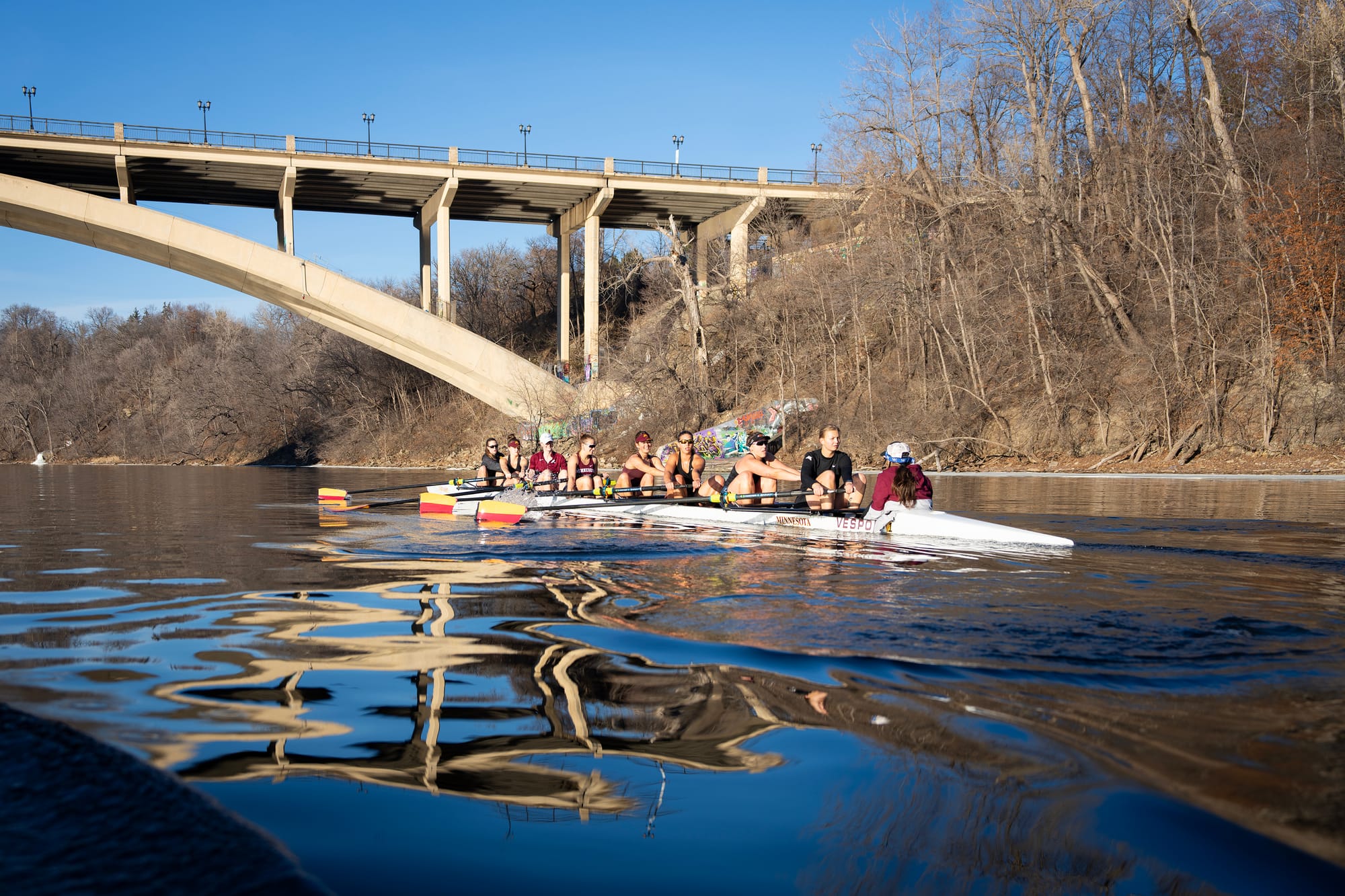 A team of women row on a boat under a bridge