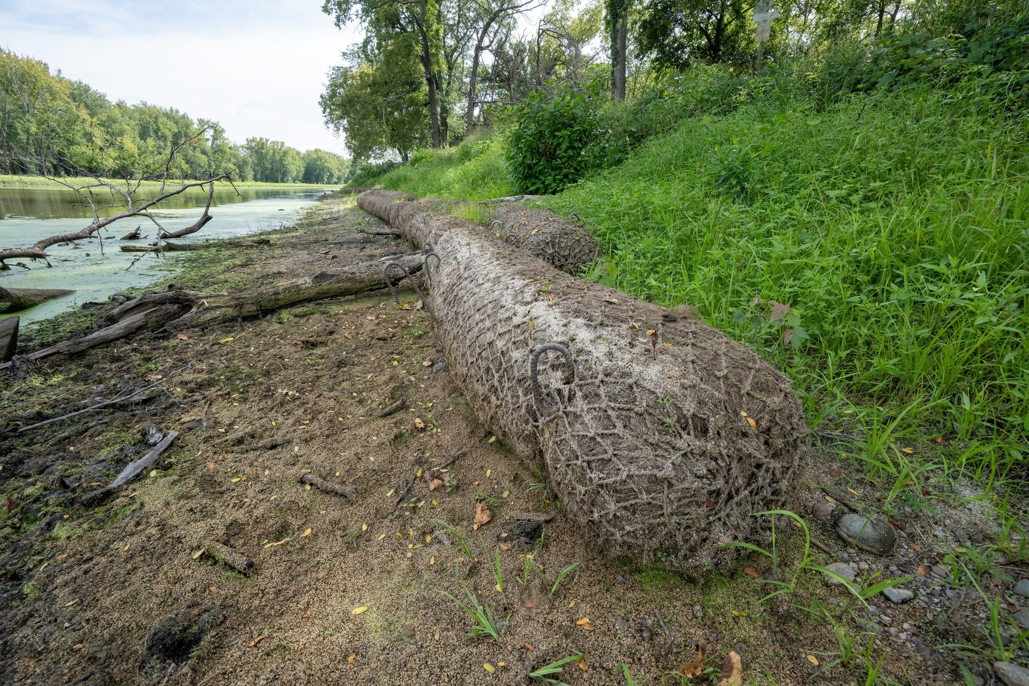 Long logs made of natural fibers are pressed up against the edge of a grassy hill. Right below the log is a short sandy bank to a river