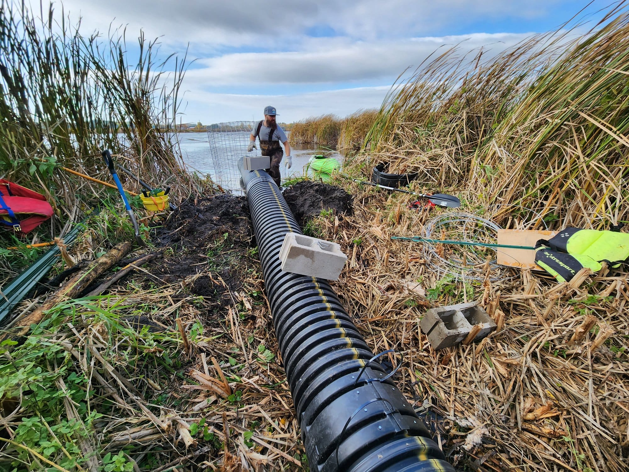 A man in camo gear works in gloves to install a long black tube into a body of water. Installation equipment surrounds it