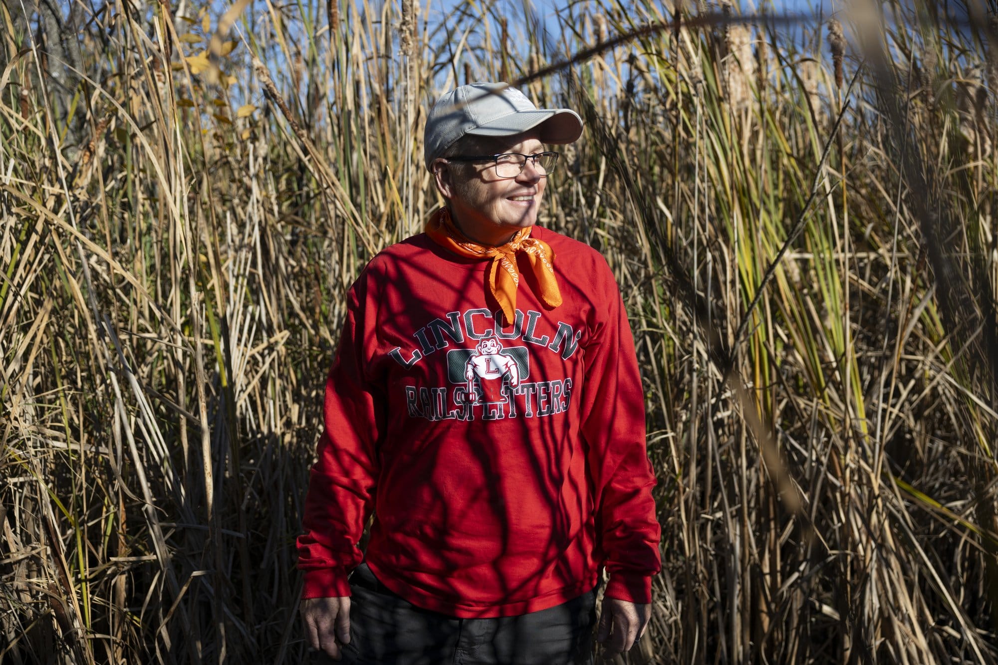 A woman in a red "Lincoln Railsplitters" school shirt, orange bandana, and ball cap stands amongst tall grass