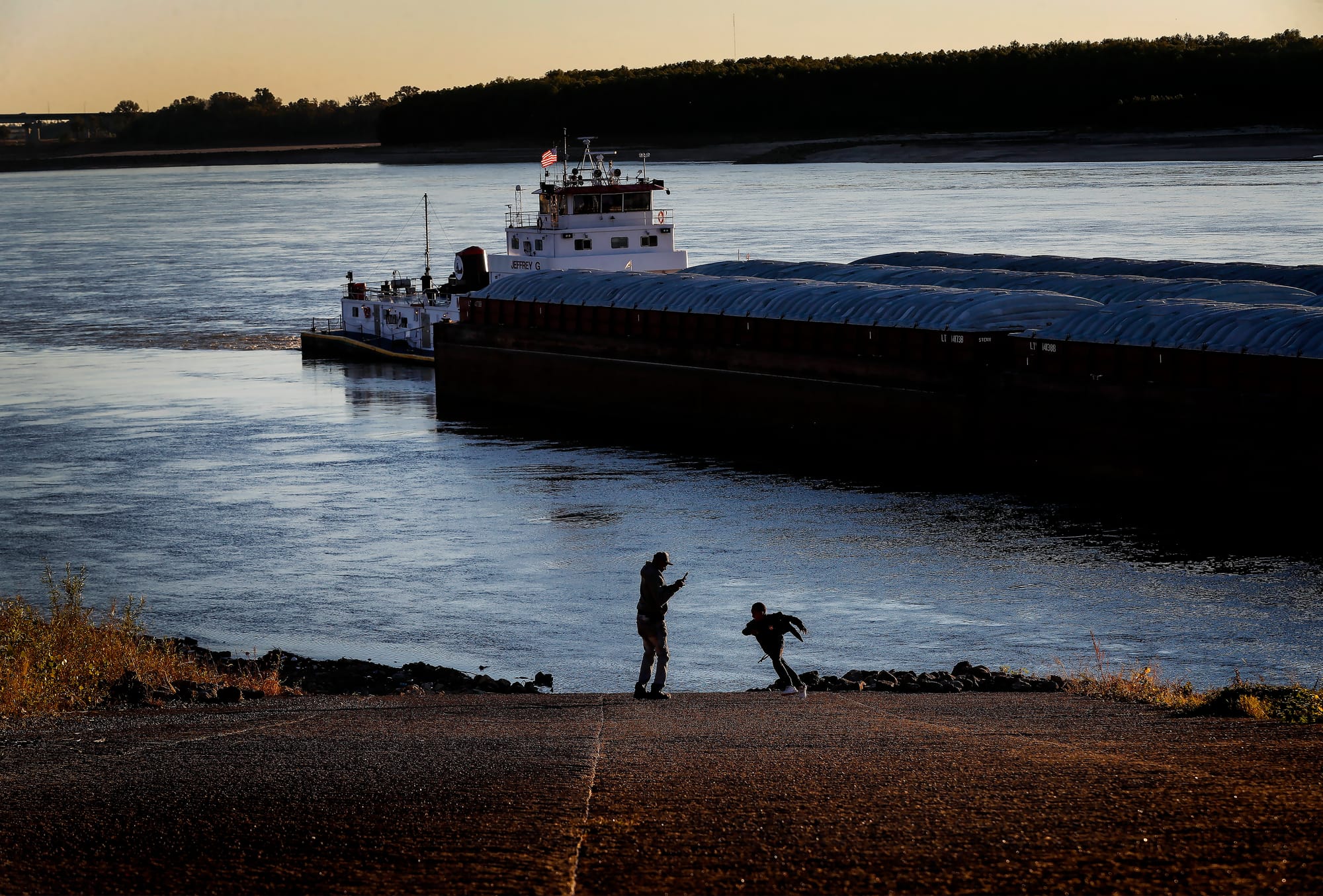 A man and child play by the river, where water is low and a barge passes by