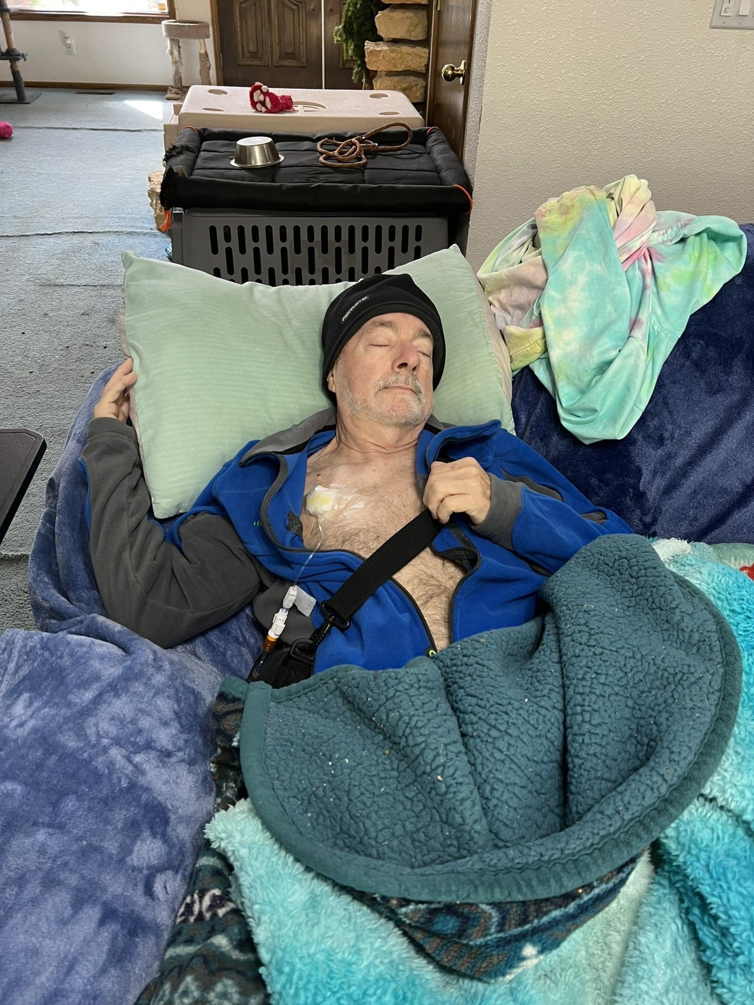 Ray Bickel sleeps in a bed covered with blankets. An IV is connected to his chest