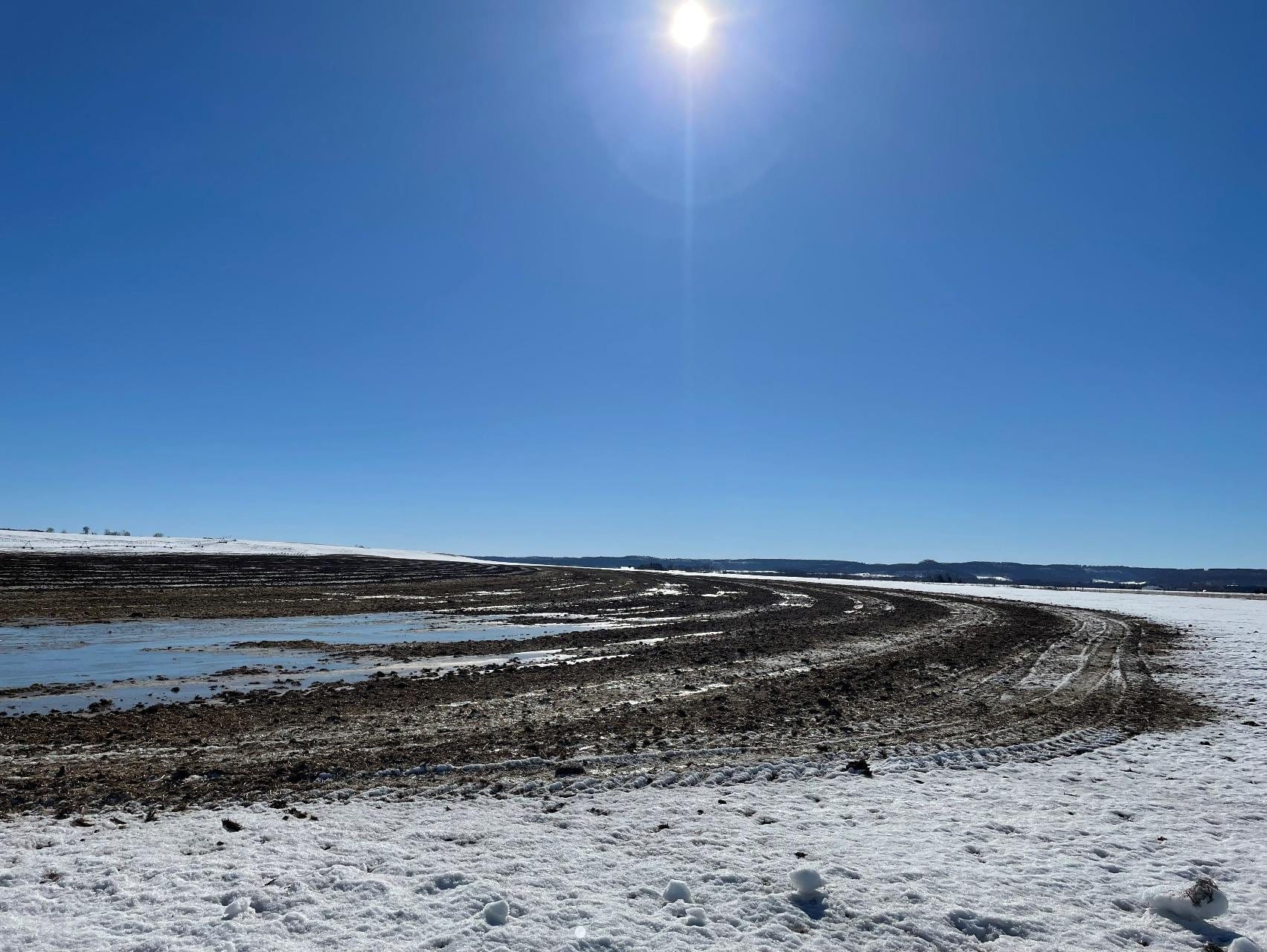 The sun shines over a snowy field covered in brown, wet, muddy tracks