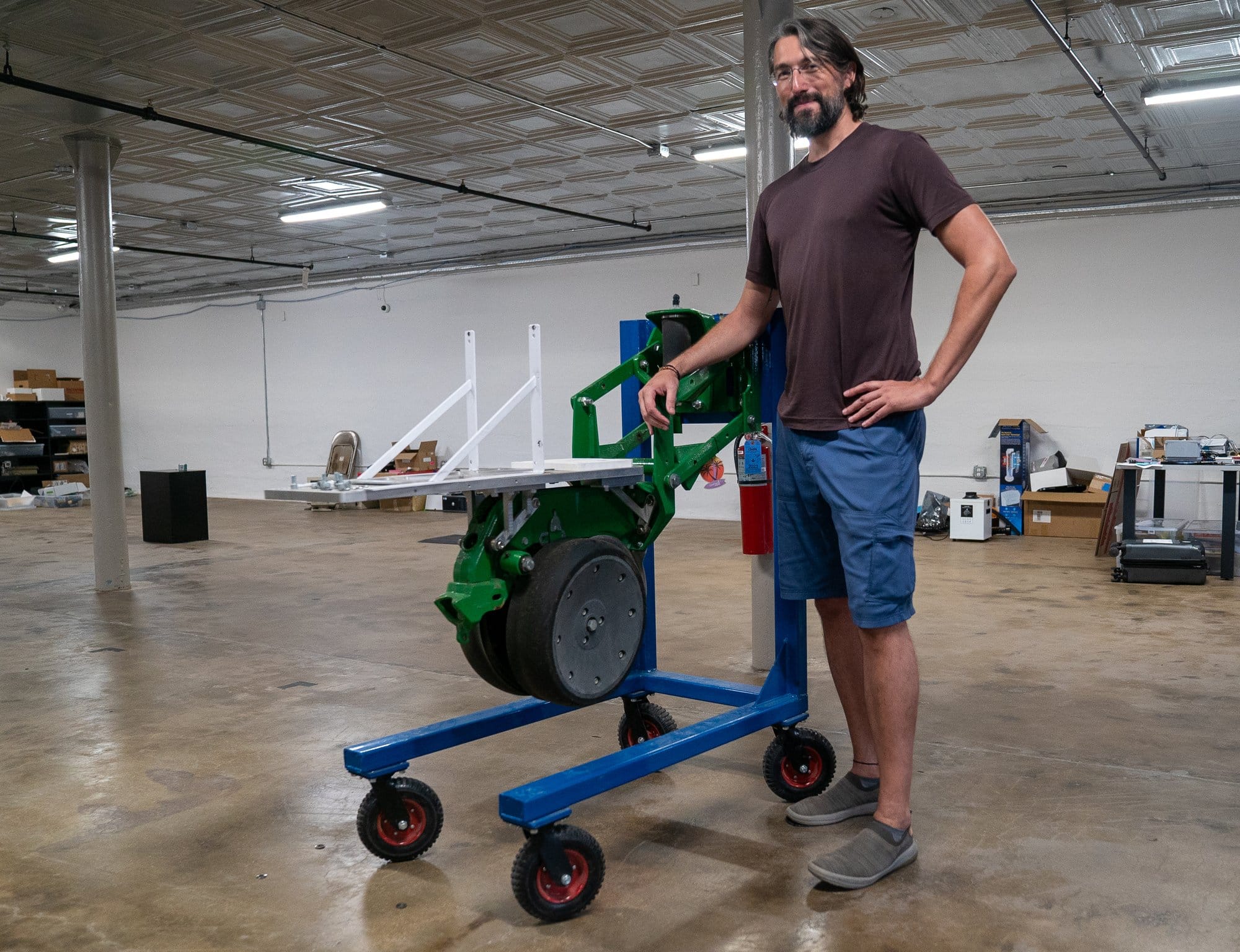 A man stands in a warehouse with a piece of farm equipment that looks like a wheel with a shelf
