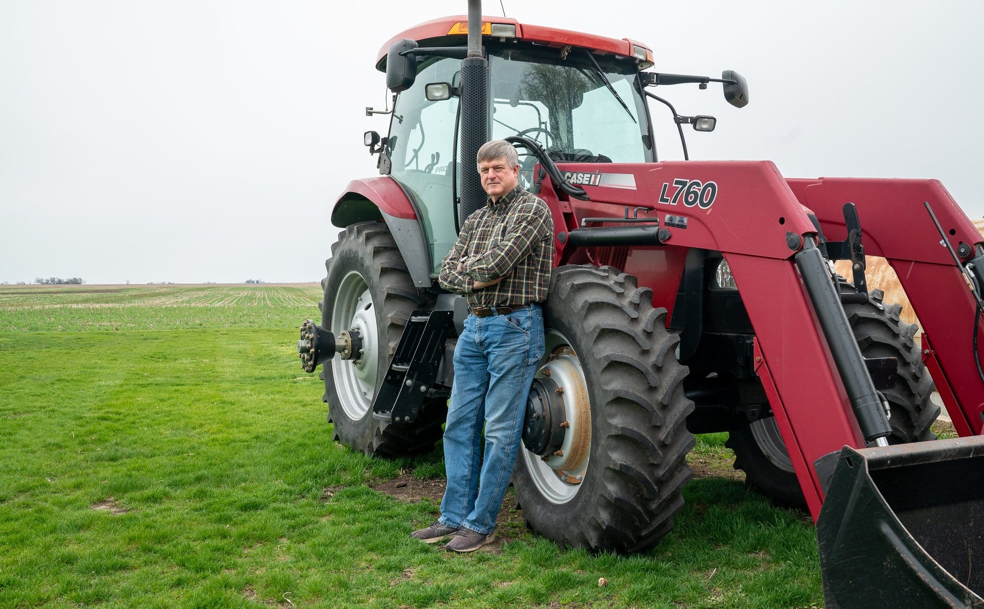 A man in a flannel and jeans stands next to a red tractor in a green field