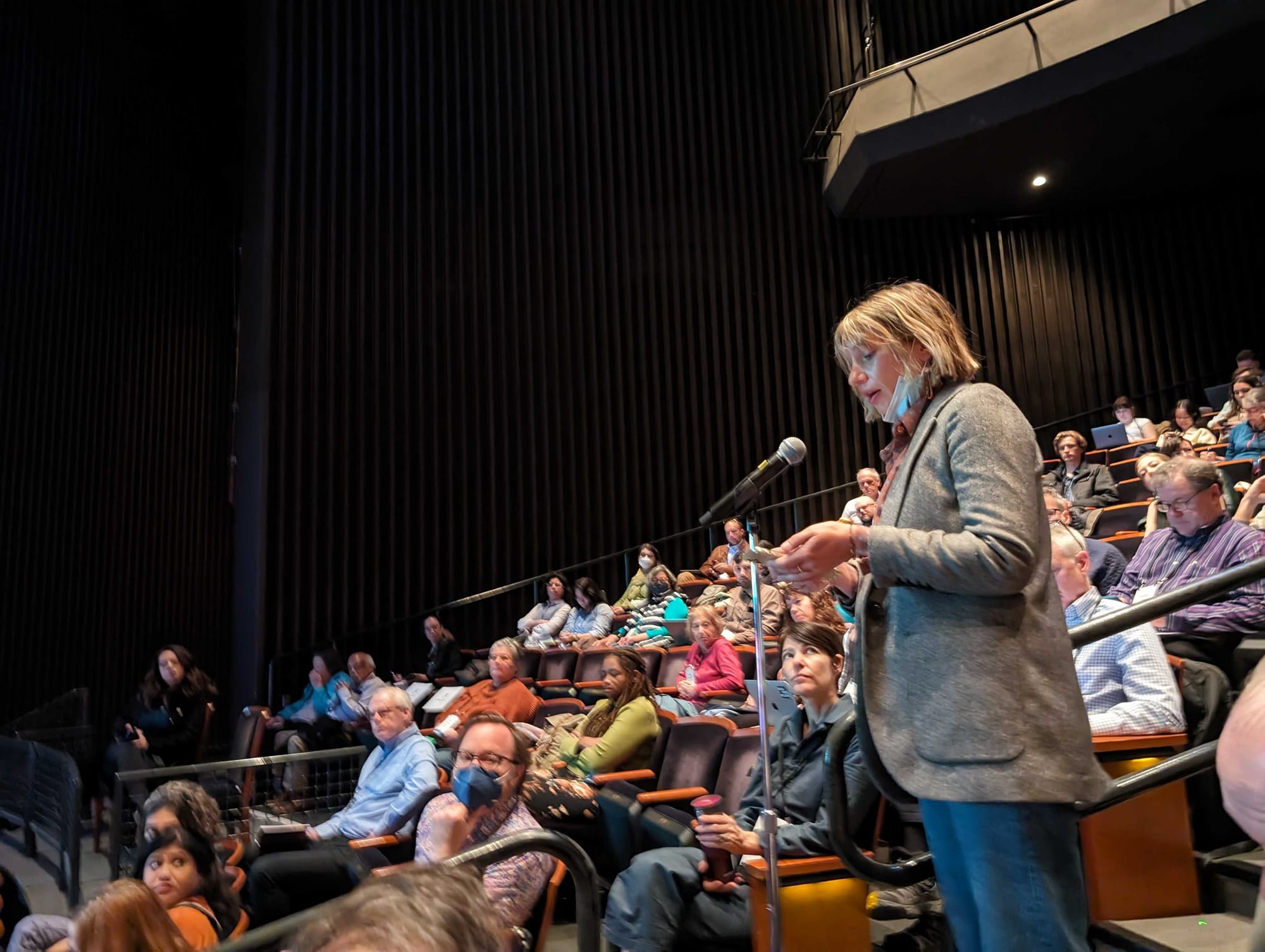 A woman stands at a mic and reads from notes in a busy conference room