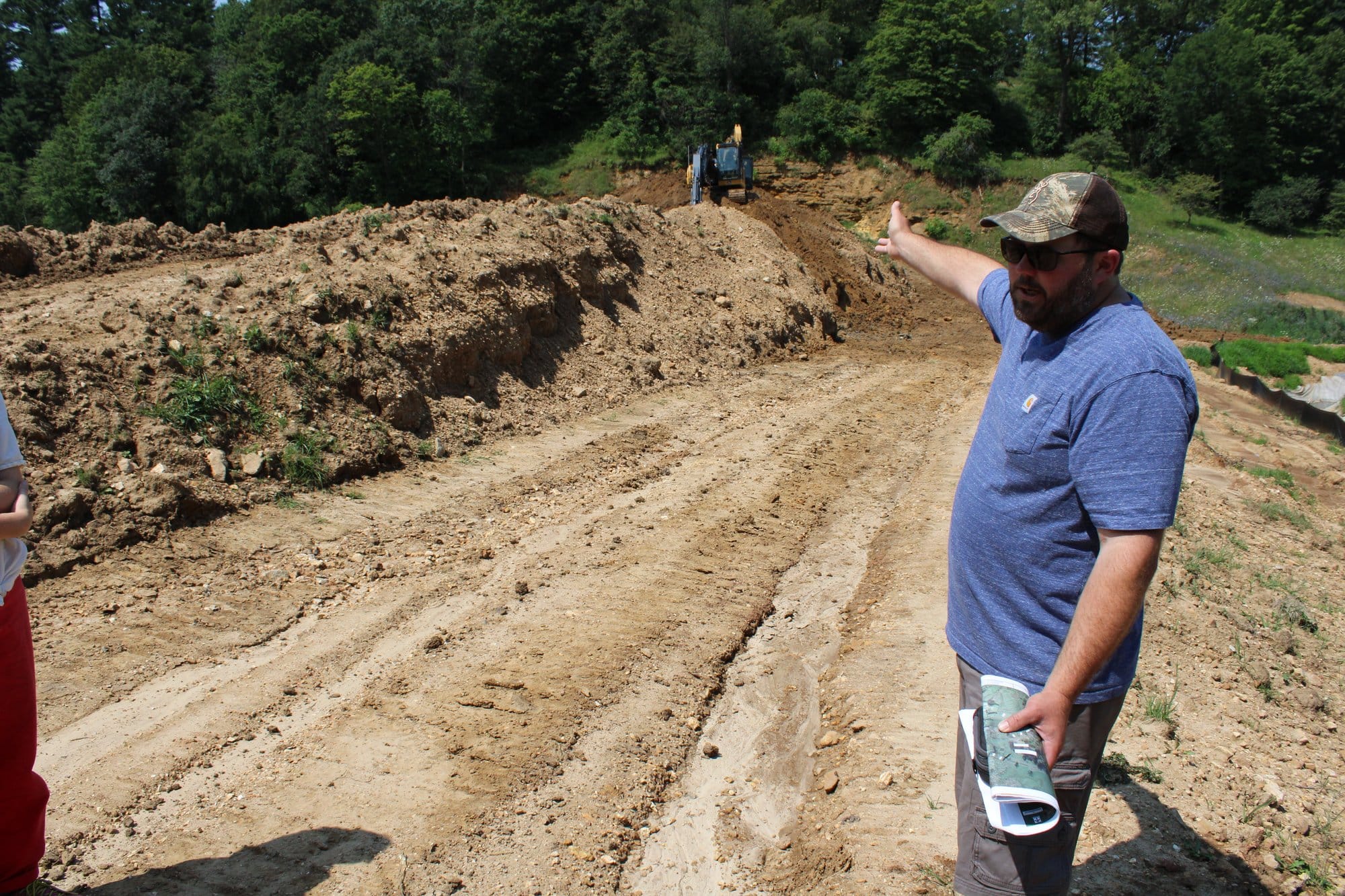 A man in a blue shirt and cap points toward excavation work at a dam site in Vernon County, Wis.