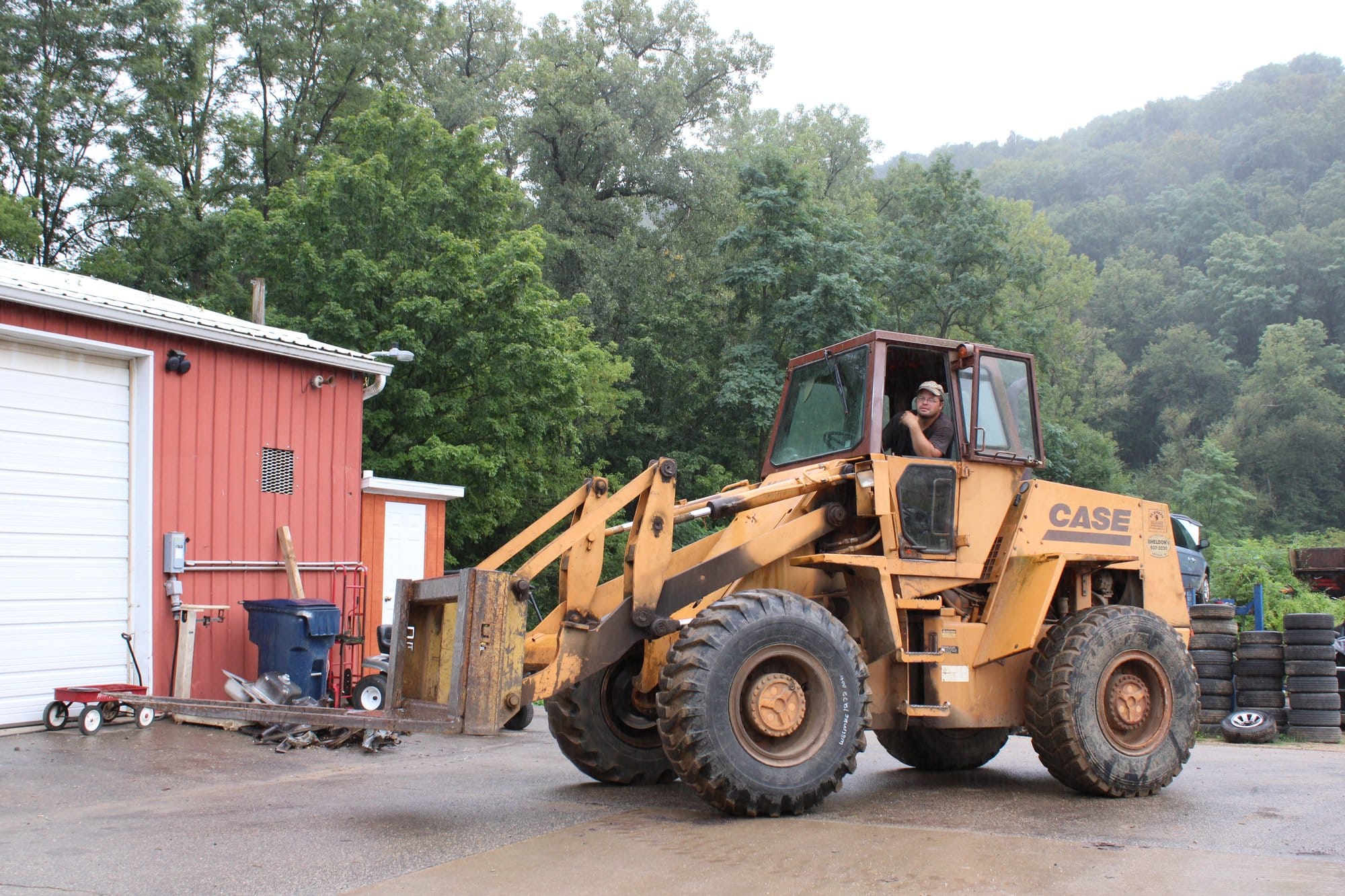A person operates a large yellow front-end loader outside a red industrial building, with trees and wooded hills in the background.