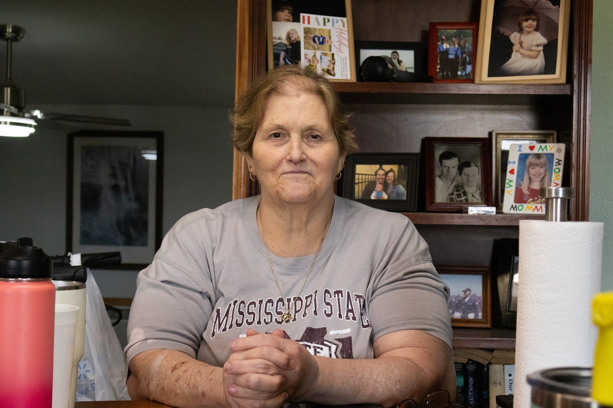 A woman in a Missisippi State shirt sits at a table in her home with framed amily photos on a shelf behind her