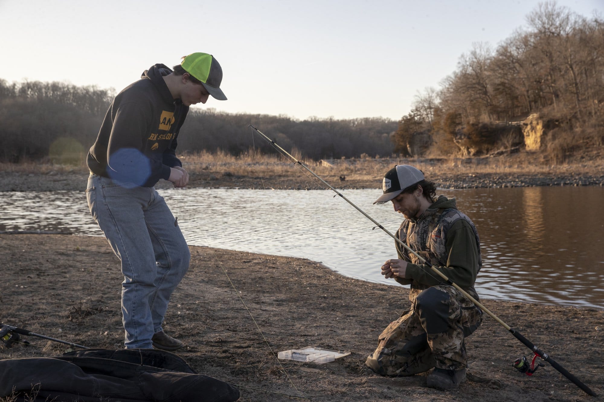 Two boys, one in camo overalls and one in jens and a hoodie, prepare fishing lines on the edge of a river