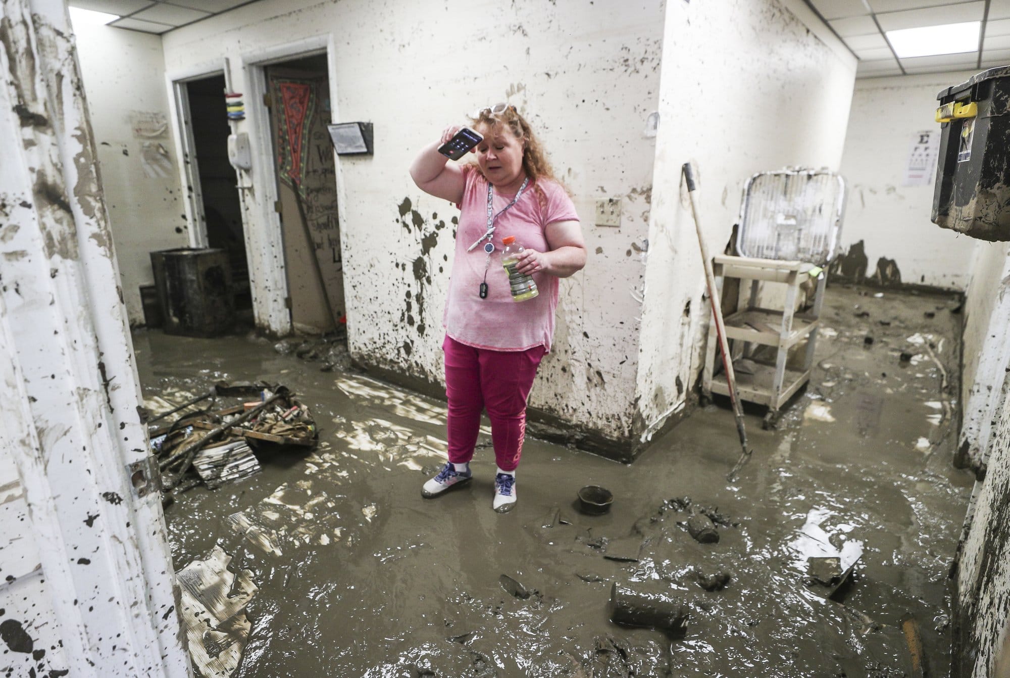A woman holds a gatorade bottle and phone in her hands. She is standing in a room where the floor and walls are covered with mud.