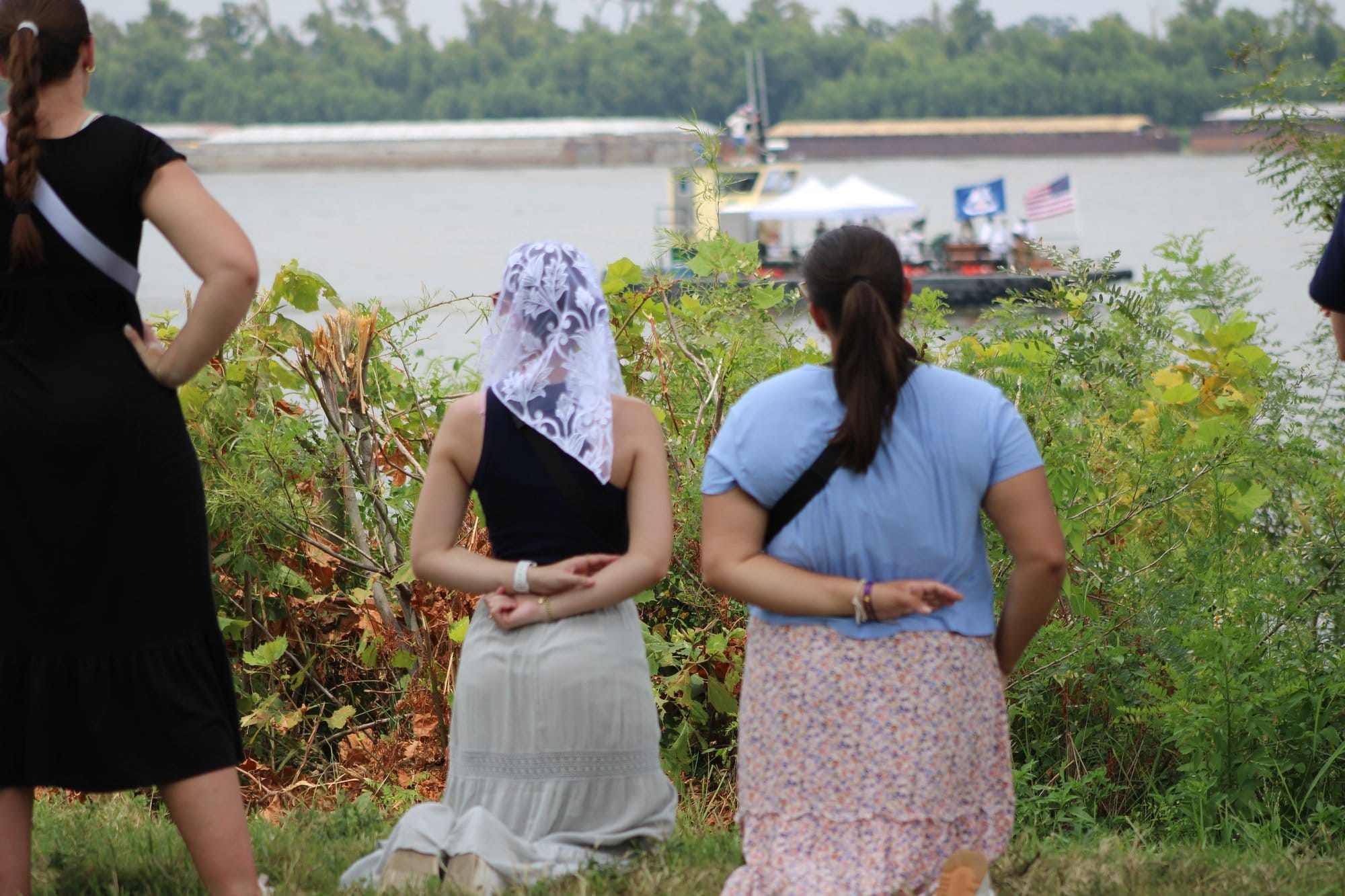 Two women kneel towards the river, where a boat is passing by. One of them dons a white lace head scarf