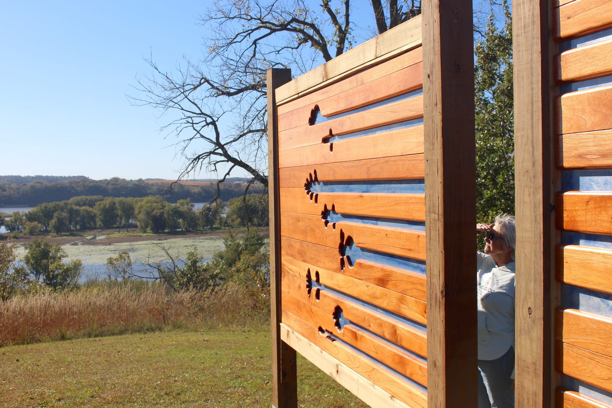 A woman stands in the sun and reads a map. On the other side of the sign, figures are carved in wood.