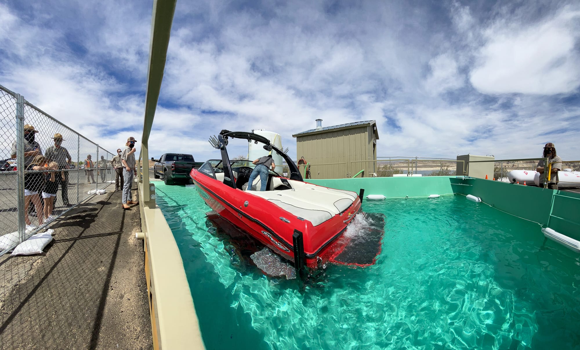 A red boat on the back of a trailer is partially submerged in a large tank containing treated water.