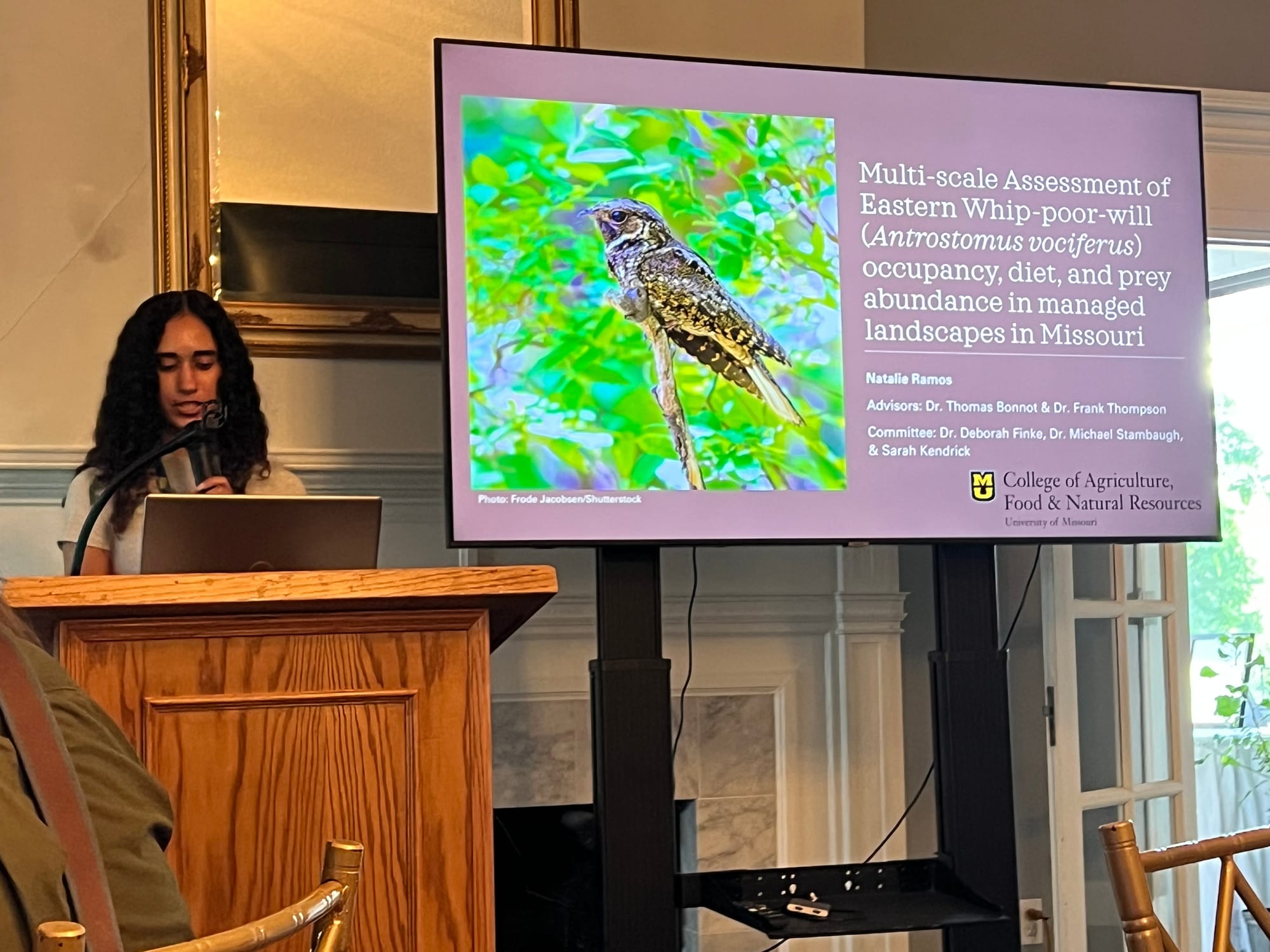 A woman stands at a podium giving a presentation. Beside her is a screen showing a whip-poor-will.