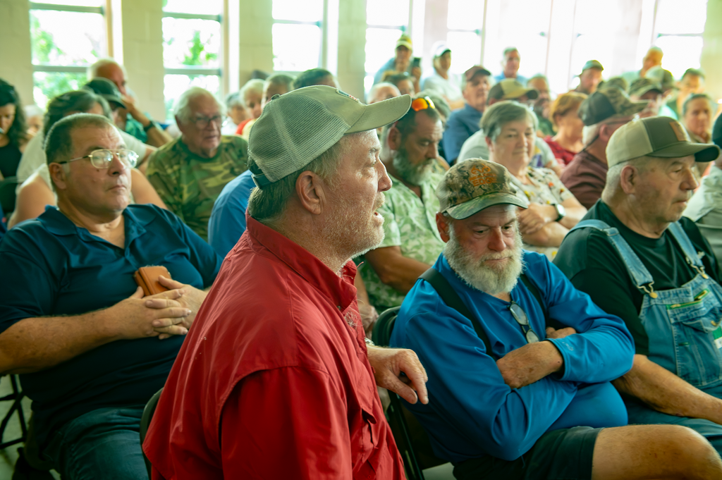 A man in a red fishing shirt and baseball cap speaks to a room full of seated, concerned-looking people.