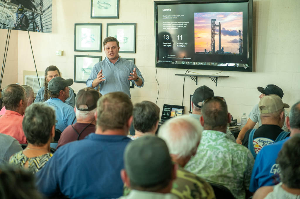 A man in a blue collared shirt stands in front of a crowded room and addresses the meeting attendants. He speaks with his hands. Starship information is displayed on a TV next to him.