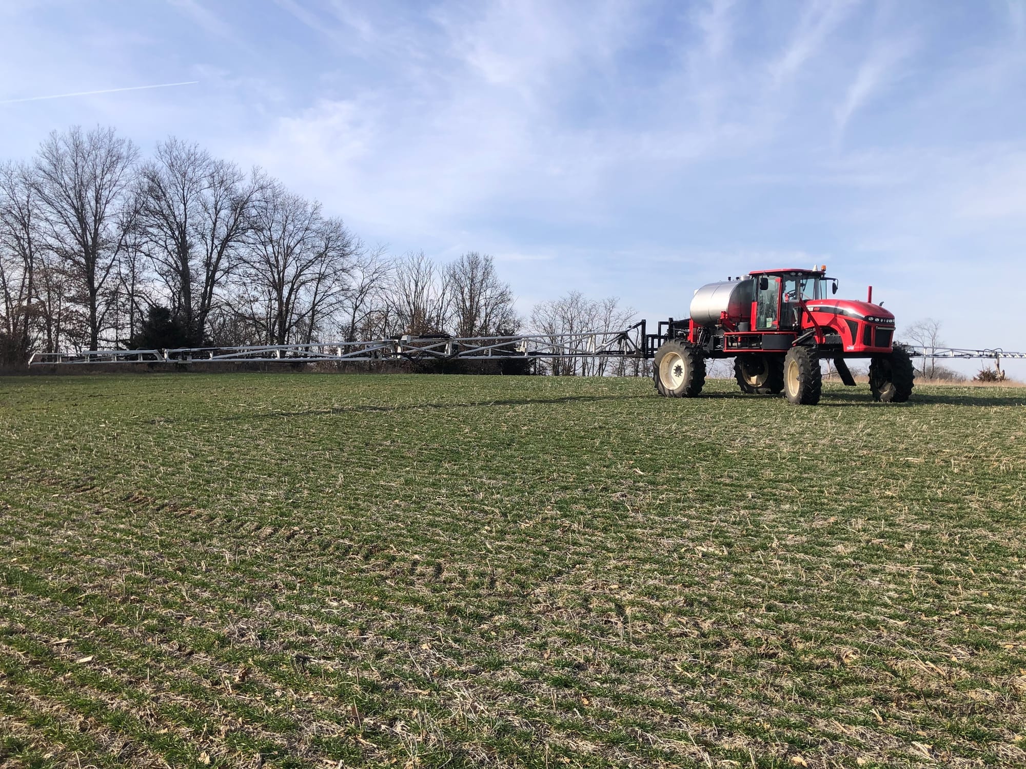 An agricultural sprayer applies fertilizer across a green farm field near Frankford, Missouri, with leafless trees in the background.
