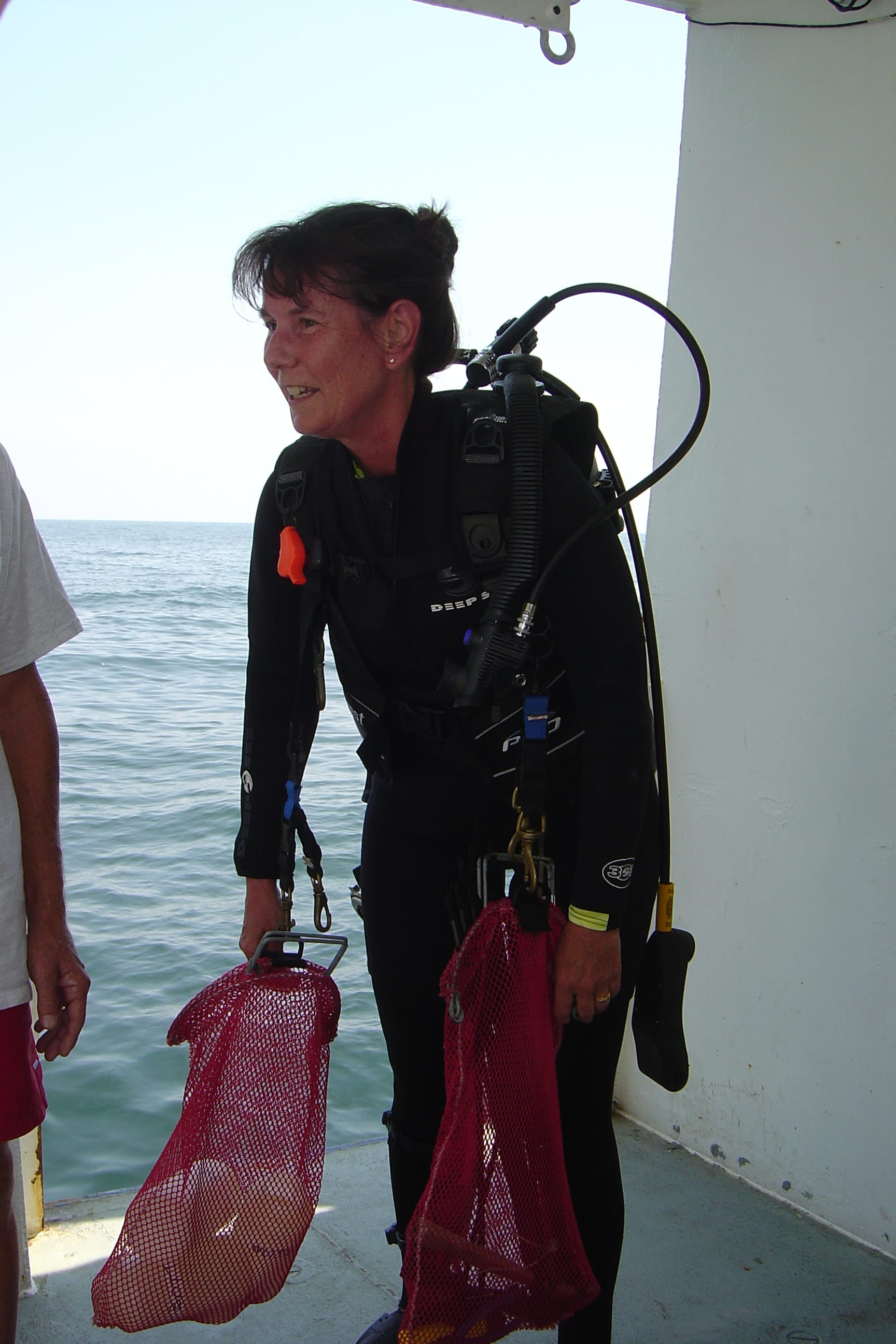Marine biologist Nancy Rabalais, wearing scuba gear, stands aboard a research vessel holding two red mesh bags with equipment, with the Gulf of Mexico in the background.