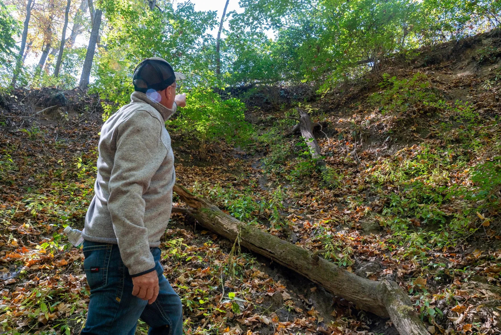 Kenny Davis stands in a wooded area, pointing to the exposed root systems where a creek bank is eroding on his property.