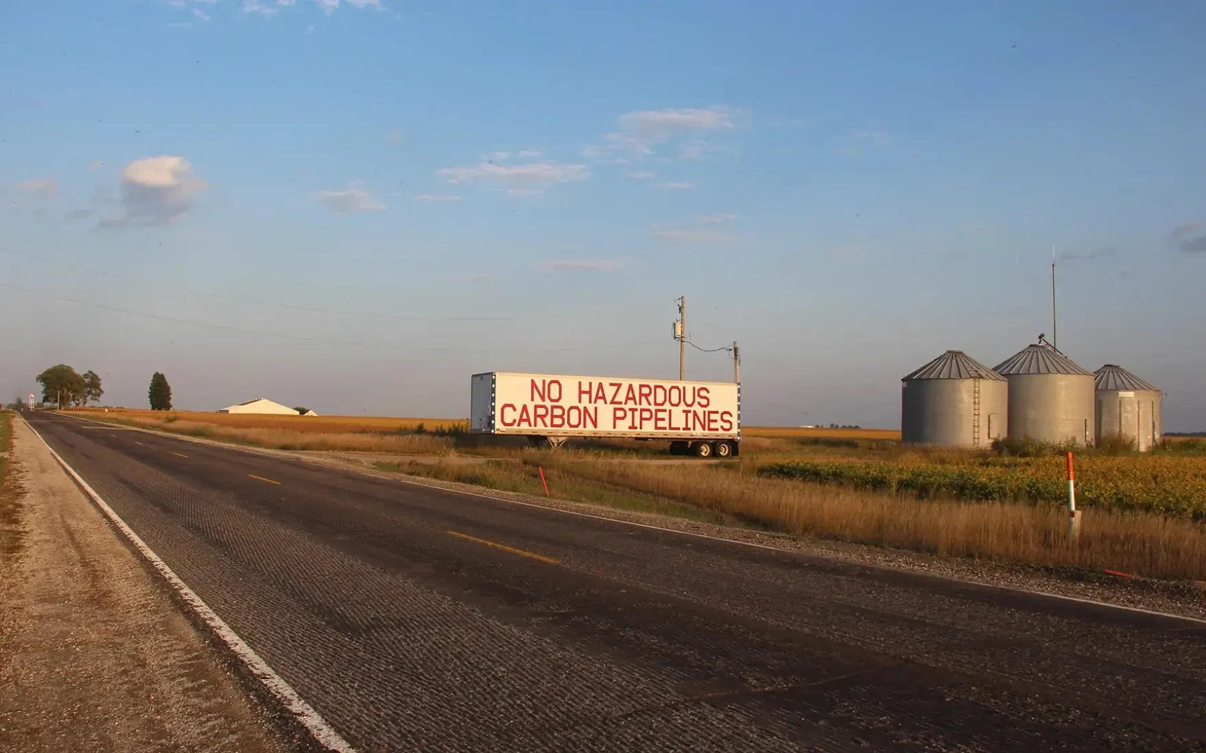 A rural Iowa road passes farmland and grain silos, with a large roadside sign reading “No Hazardous Carbon Pipelines.”