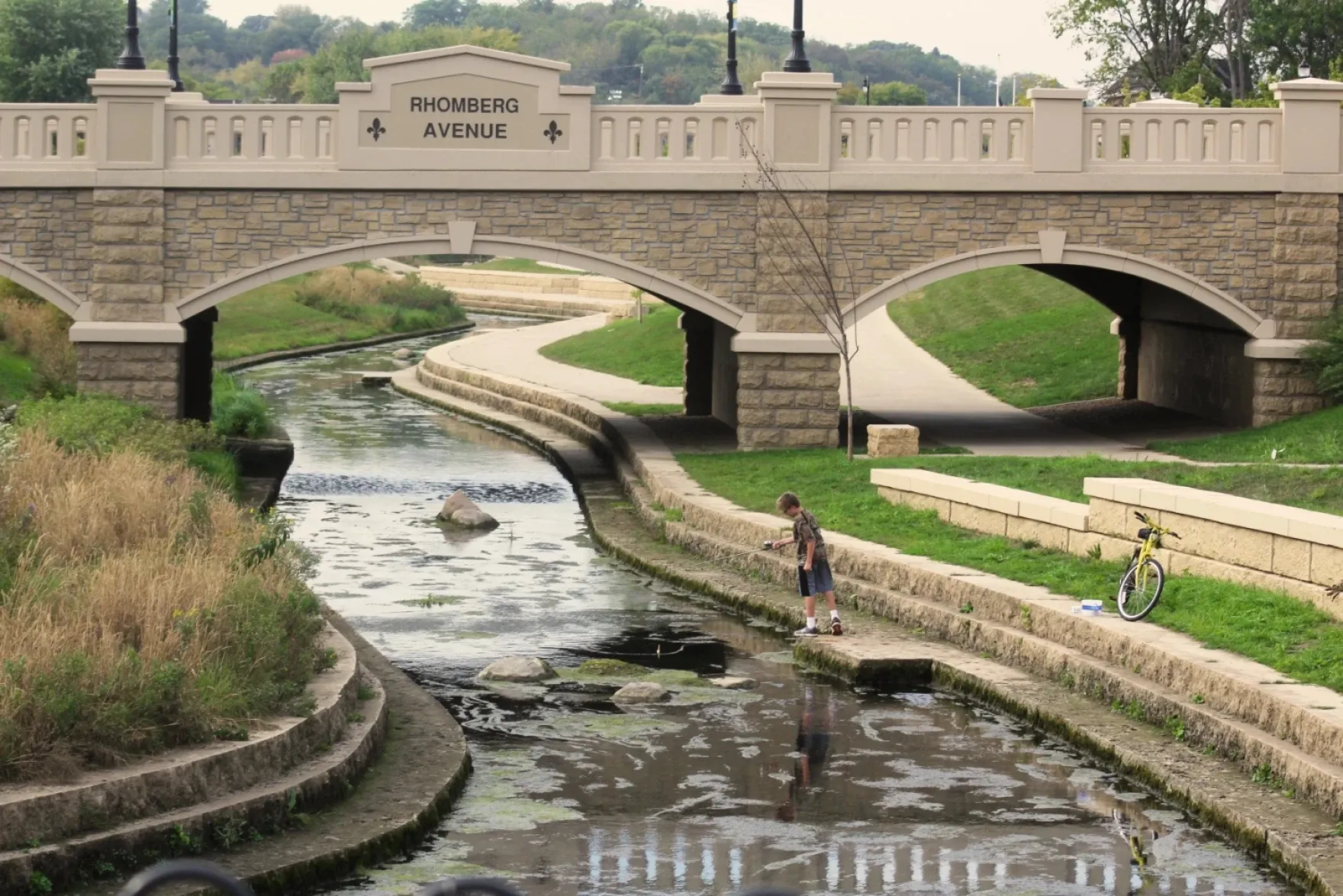 A child fishes in Bee Branch Creek in Dubuque, Iowa, beside a bicycle and walking path, with the stone Rhomberg Avenue bridge spanning the creek.