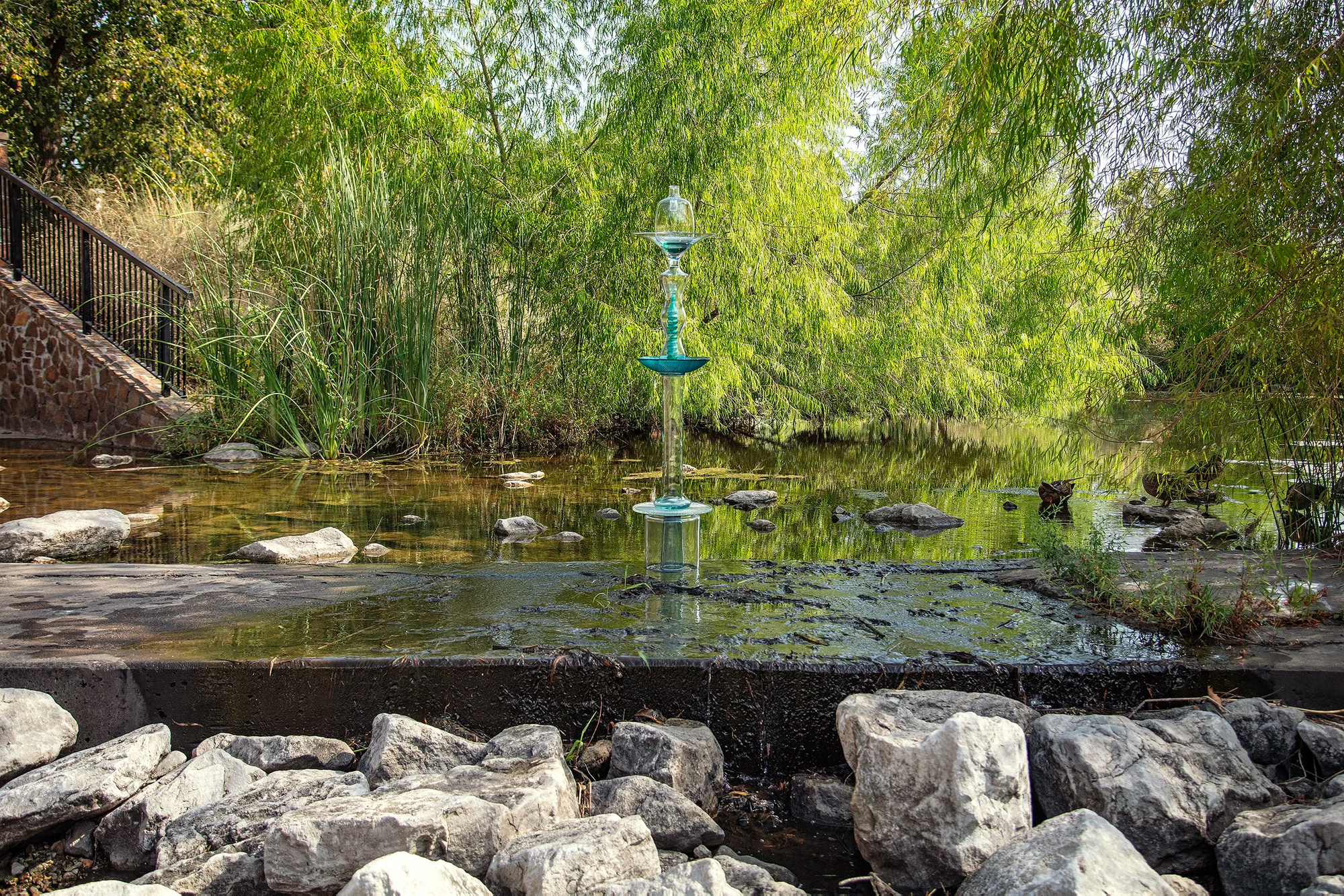 A blue-green glass sculpture stands tall in a quiet creek surrounded by dense vegetation, rocks and willow trees.