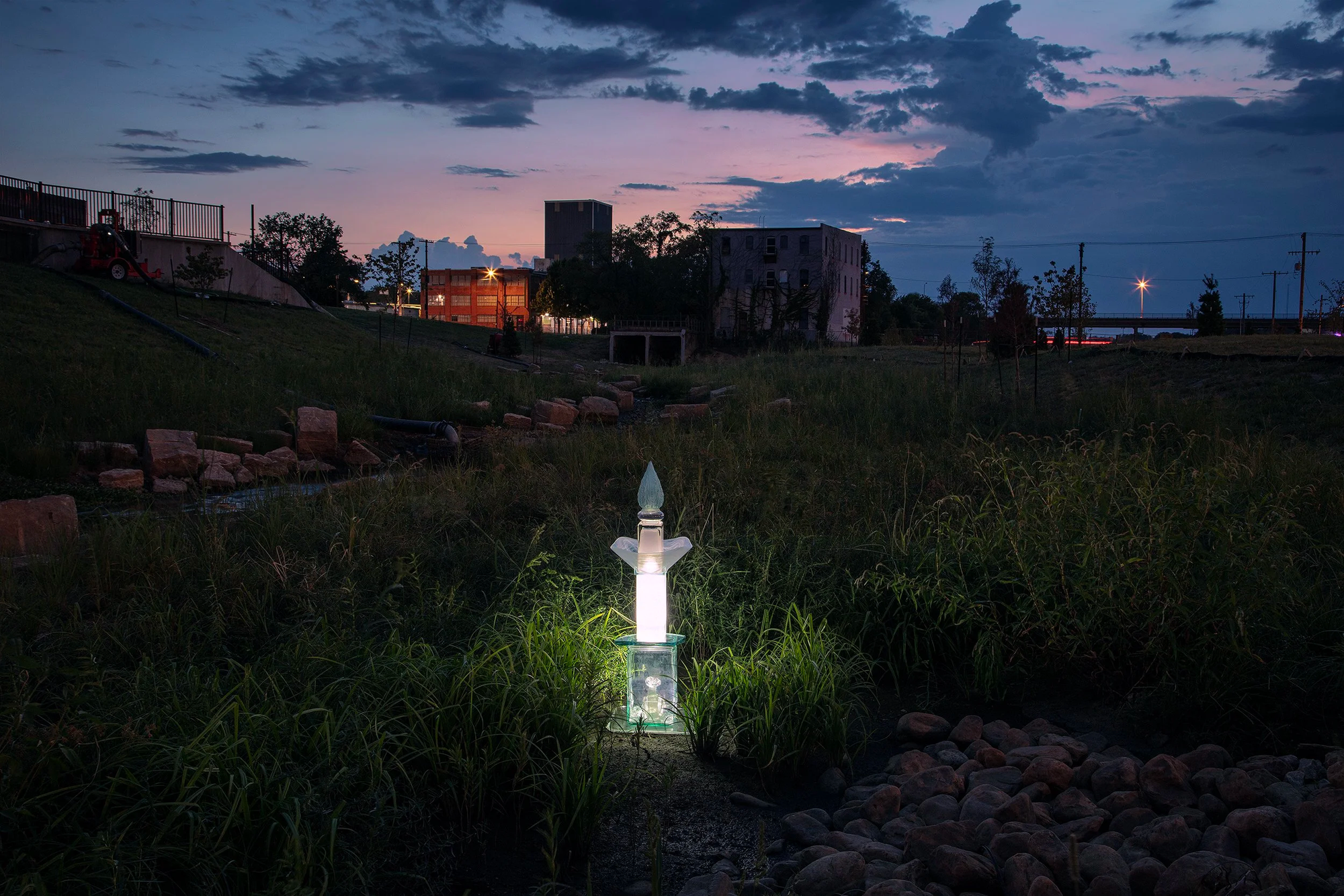 A glass sculpture glows ghostly white at dusk. A dark silhouette of a building rises in the background.