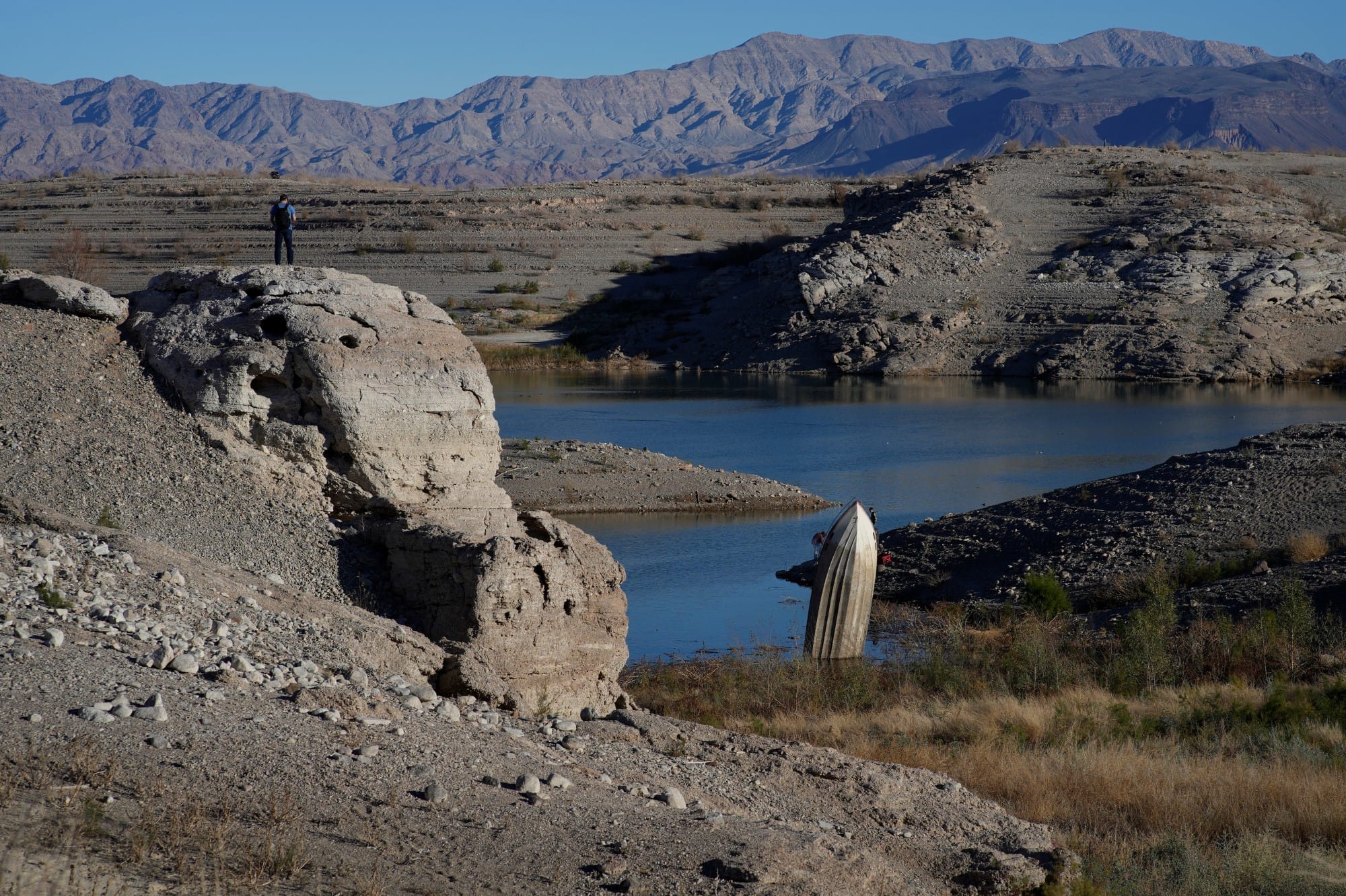 A man stands on a hill overlooking a formerly sunken boat standing upright into the air with its stern buried in the mud along the shoreline of Lake Mead at the Lake Mead National Recreation Area.