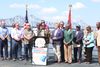 A Black woman speaks into a mic at a podium surrounded by other mayors beside the river. A bridge rises crosses the river behind them.