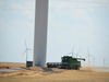 A tractor moves over a field of yellow wheat. Nine wind turbines are seen
