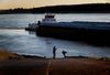 A man and child play by the river, where water is low and a barge passes by