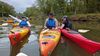 Several people gather together in their kayaks on the river. One person points to something off camera
