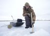 A man in winter gear kneels behind a hole in a large sheet of ice. He lowers a measuring stick into the hole, and behind him are several pieces of sceince equipment