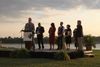 A group of local officials stands on an outdoor stage beside the Mississippi River as a man speaks at a podium during a mayors' meeting.